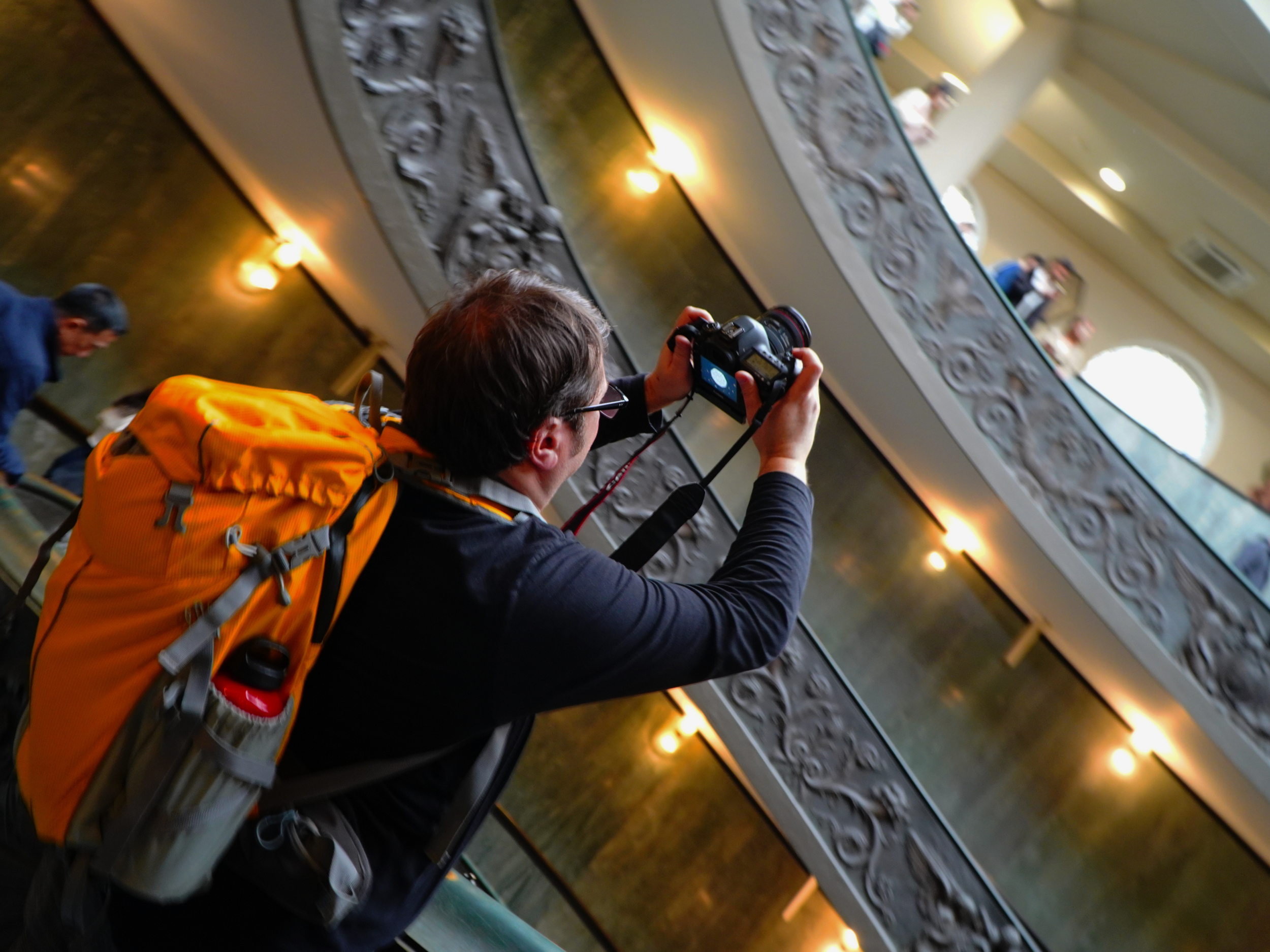 Une personne avec un sac à dos orange prend une photo à l'intérieur d'un bâtiment avec une décoration métallique et des personnes au balcon.