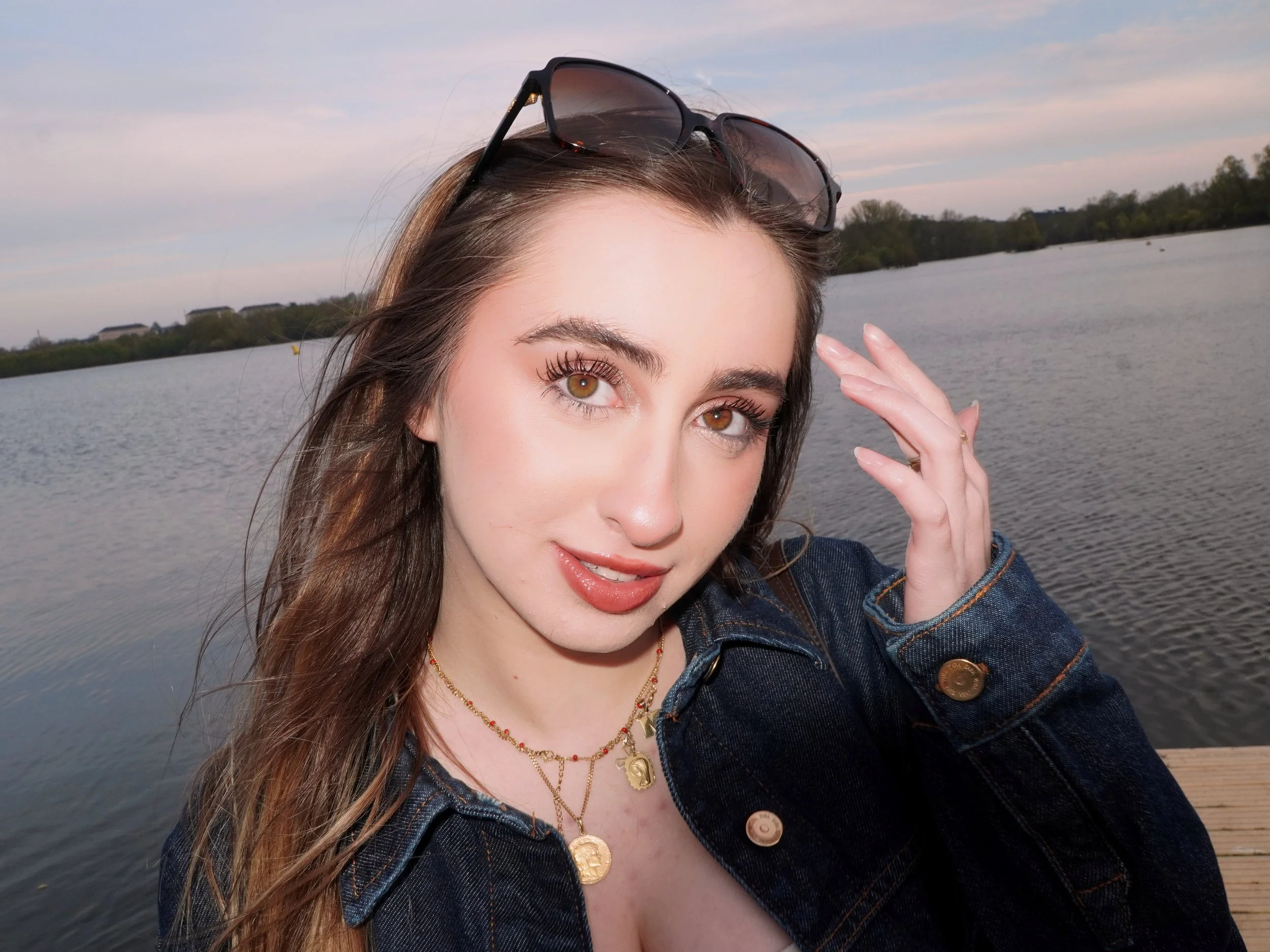 Jeune femme aux cheveux longs, portant des lunettes sur la tête, souriant à la caméra au bord d'un lac lors du coucher du soleil.