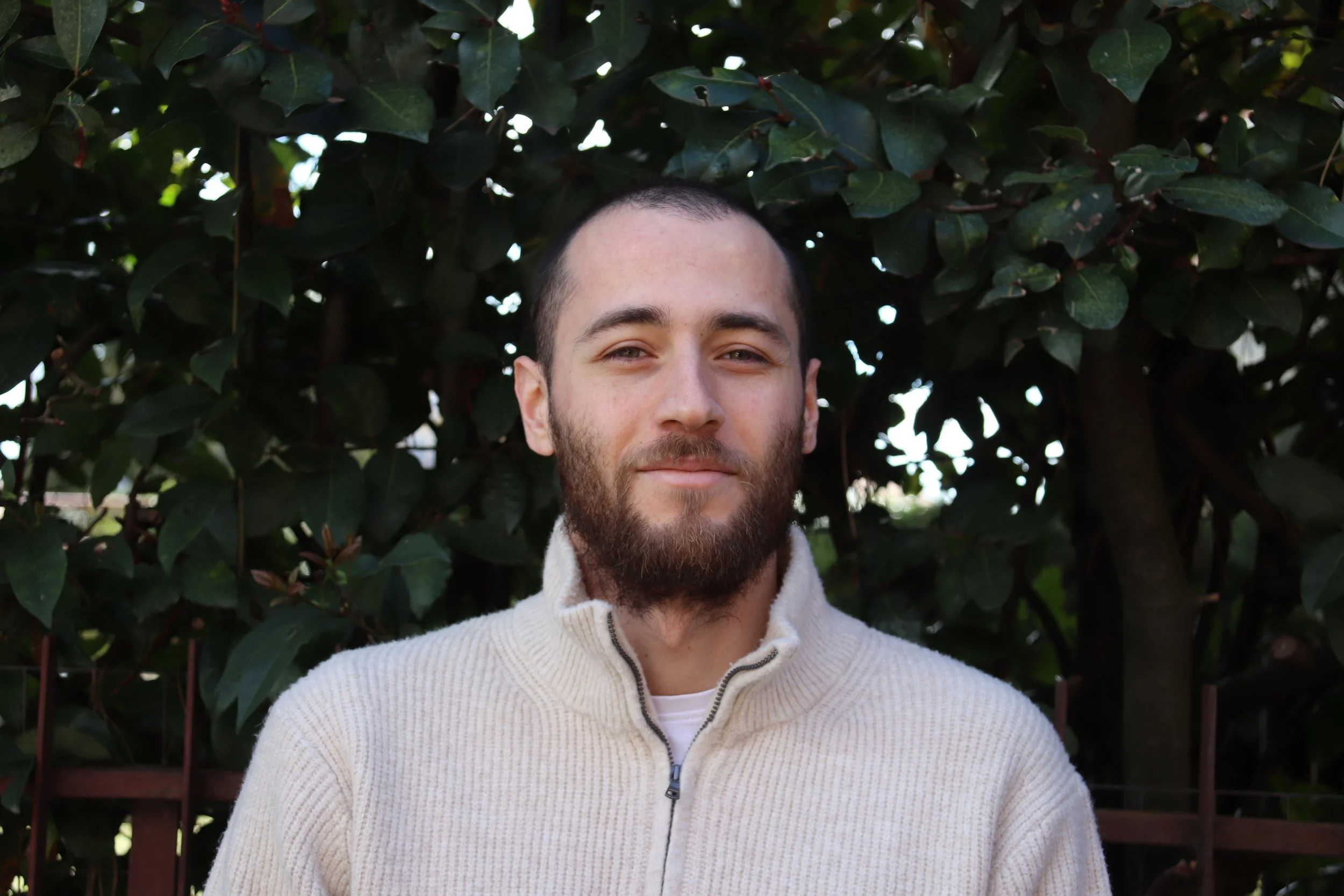 A man with a beard and short hair, smiling slightly, wearing a cream-colored zip-up sweater, standing outdoors in front of dark green leafy bushes.