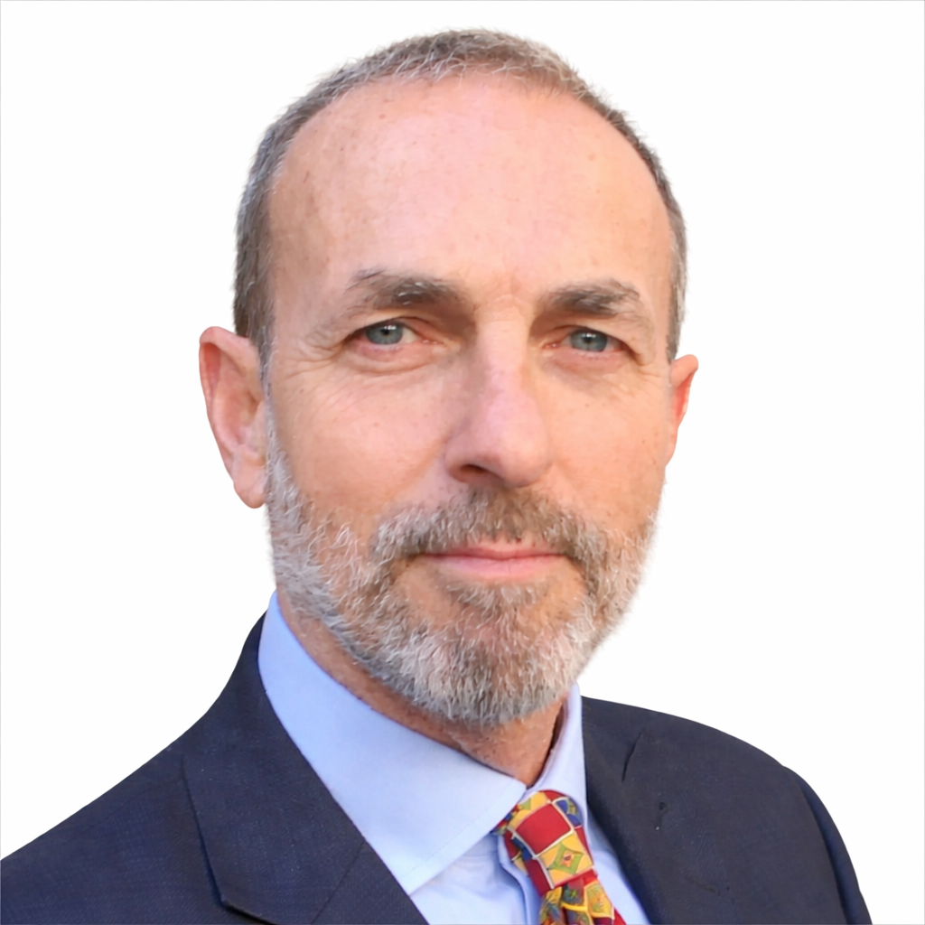 Close-up portrait of a middle-aged man with a beard and short hair, wearing a dark suit, light blue shirt, and patterned tie, against a plain white background.