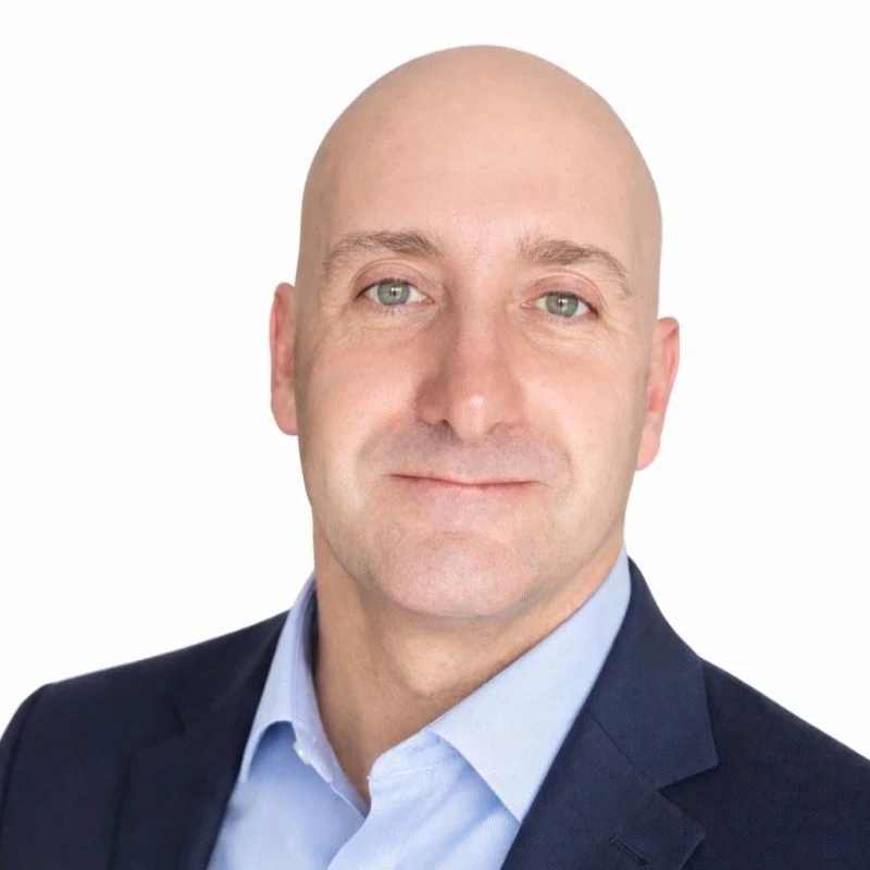 Headshot of a bald man wearing a suit and light blue shirt, smiling slightly against a white background.