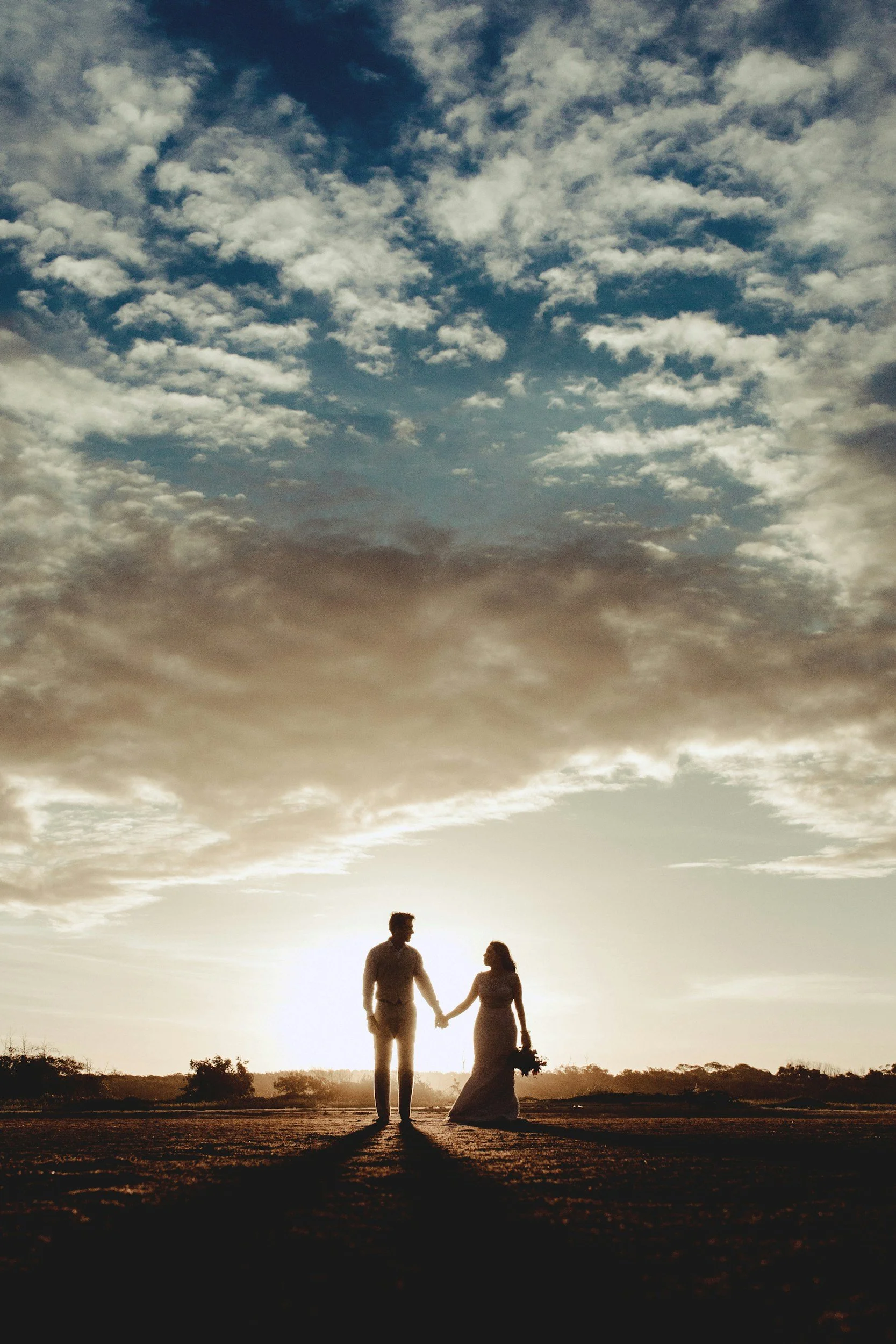 A man and a woman holding hands and walking on a pathway at sunset with a cloudy sky overhead.