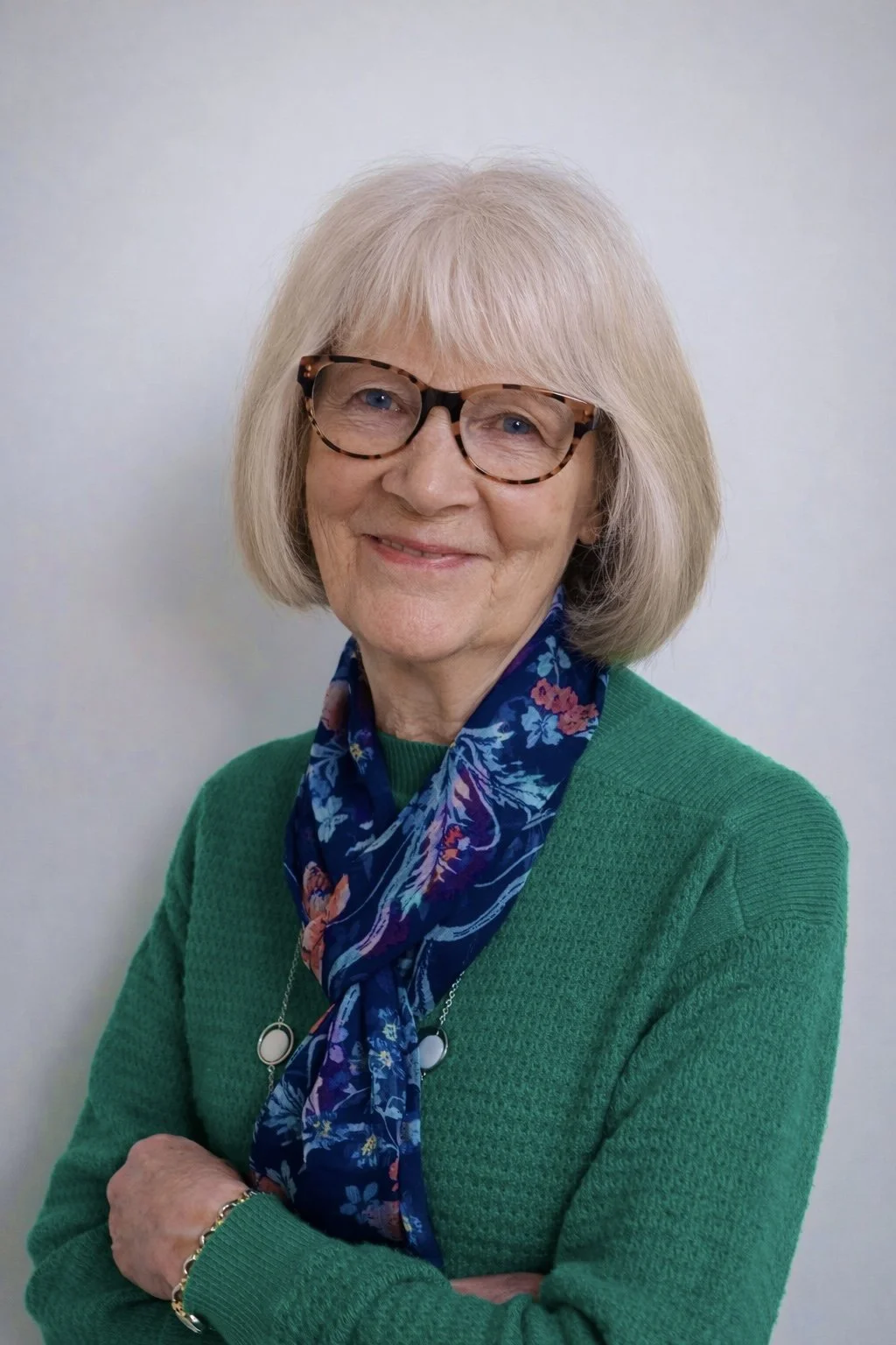 A smiling elderly woman with glasses, wearing a green sweater, a blue floral scarf, and a necklace, stands against a plain light background.