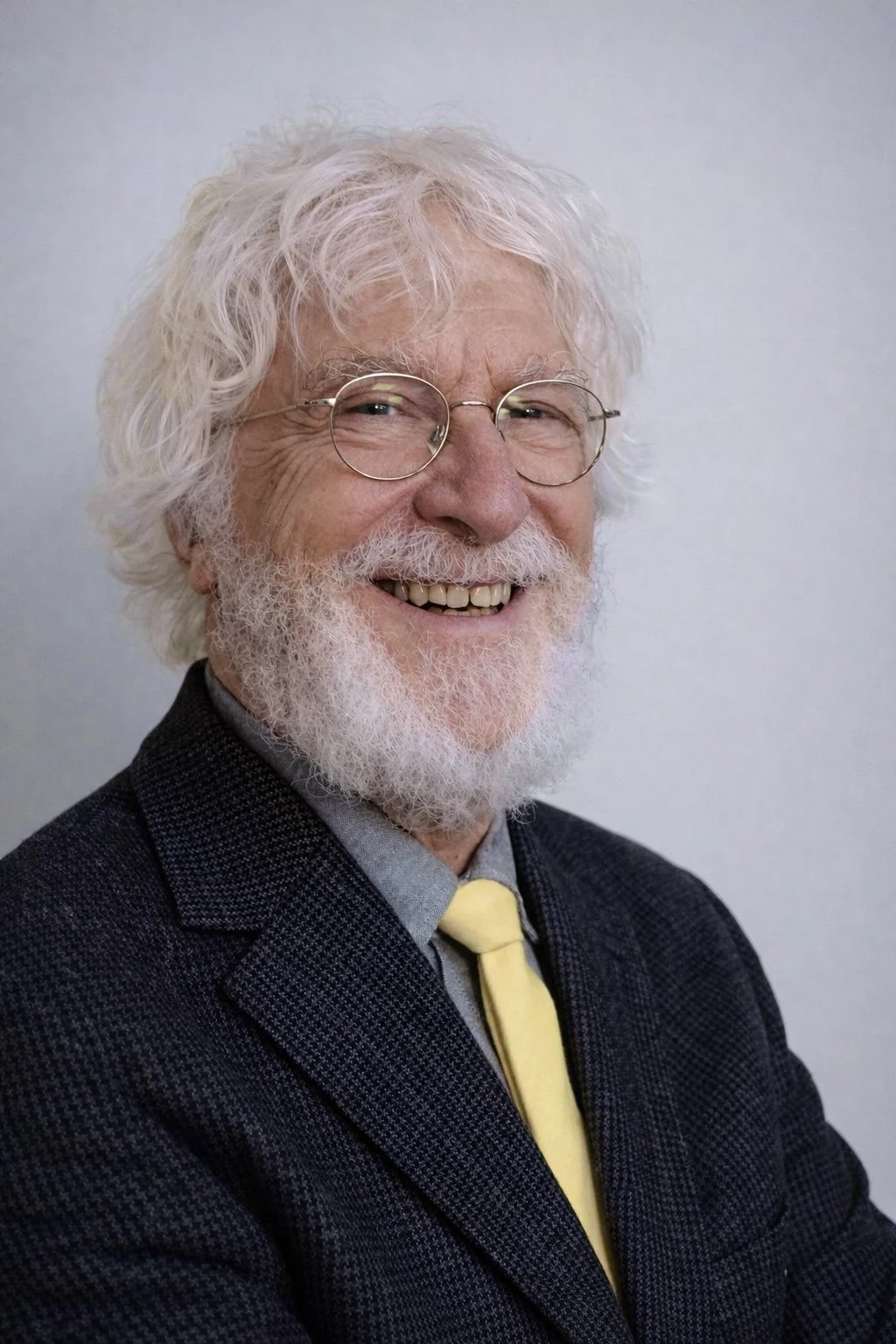 Portrait of an elderly man with curly white hair, a full white beard, round glasses, smiling, wearing a dark blazer, light gray shirt, and yellow tie, against a plain light background.