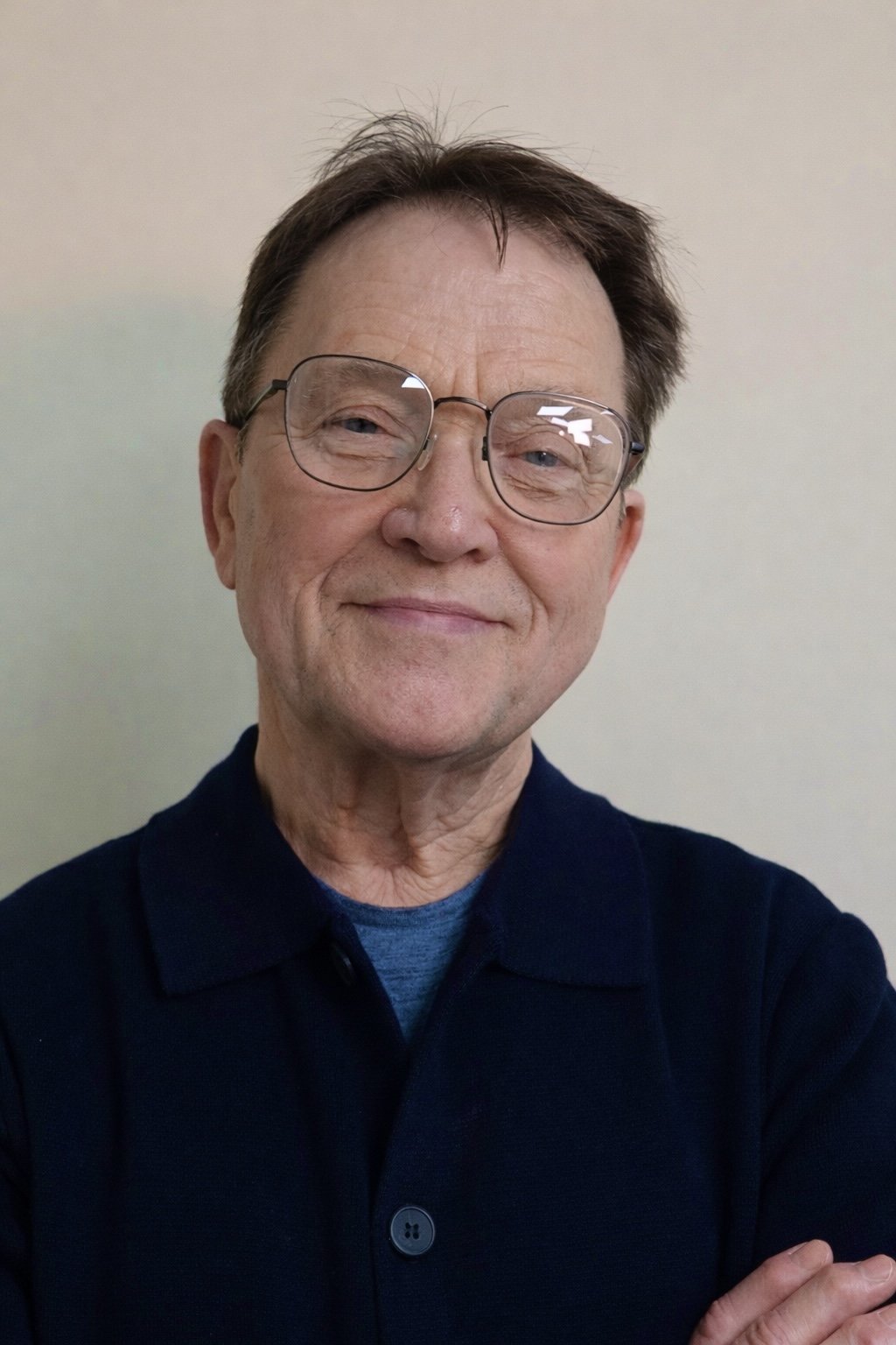 A middle-aged man with glasses smiling, wearing a dark collared shirt, standing in front of a plain light-colored wall.