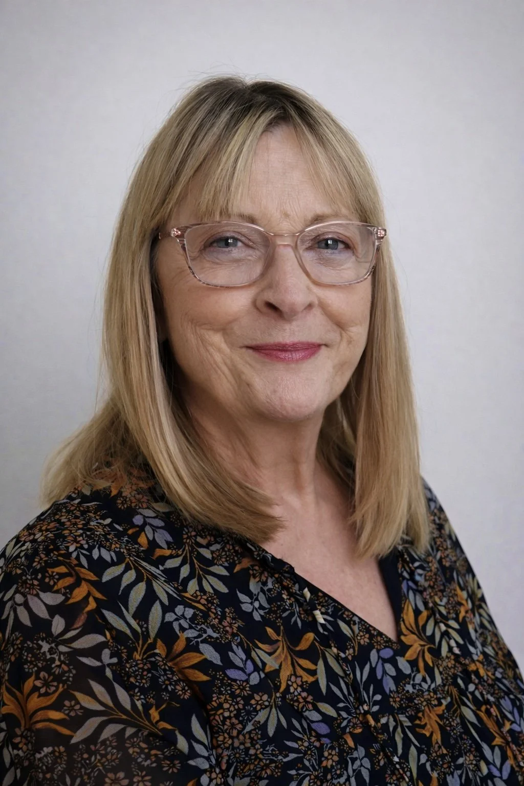 A middle-aged woman with blonde hair, glasses, and a floral patterned blouse, smiling in front of a plain white background.