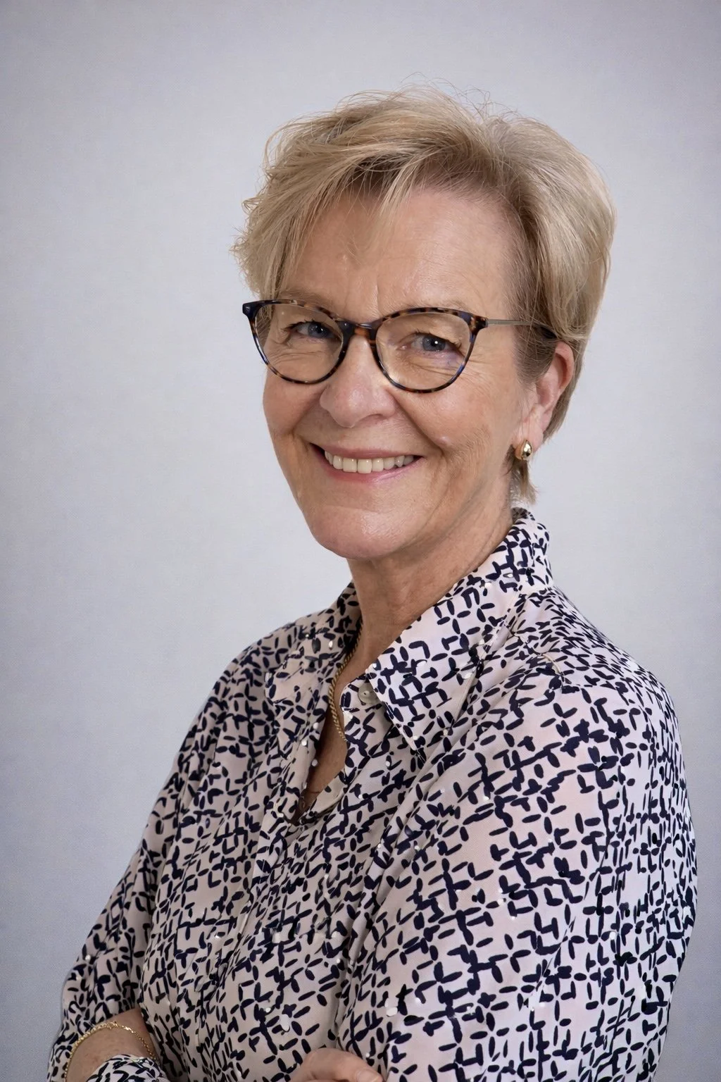 A smiling middle-aged woman with short blond hair, wearing glasses, gold earrings, a patterned blouse, and a gold bracelet, standing against a plain gray background.