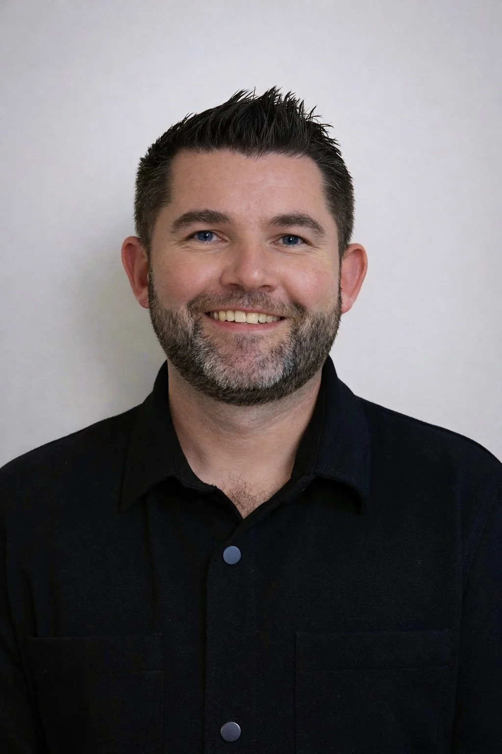 Headshot of a smiling man with short dark hair, a beard with some gray, and blue eyes, wearing a black shirt against a plain light-colored background.