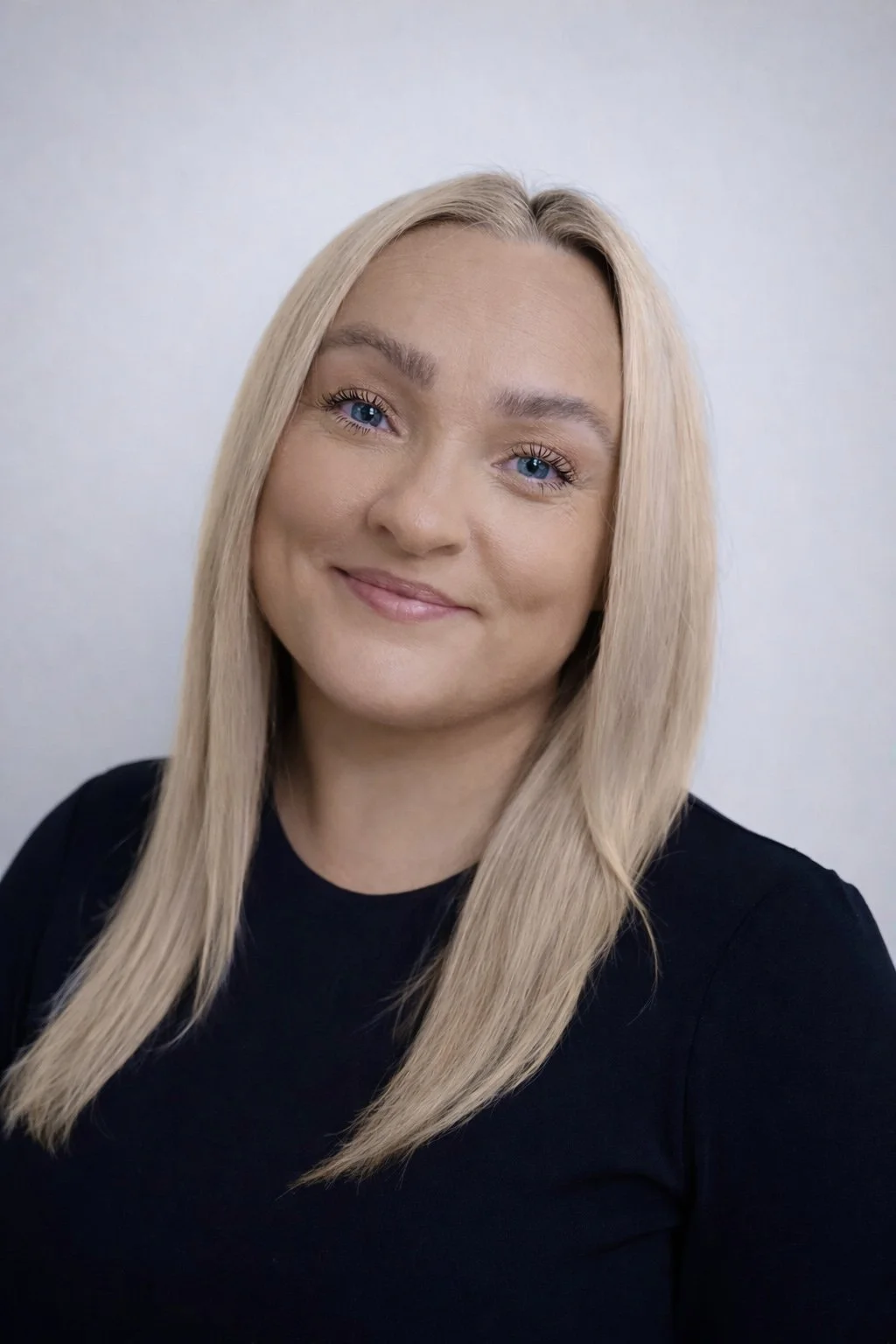 A woman with long blonde hair and blue eyes smiling at the camera, wearing a black top.