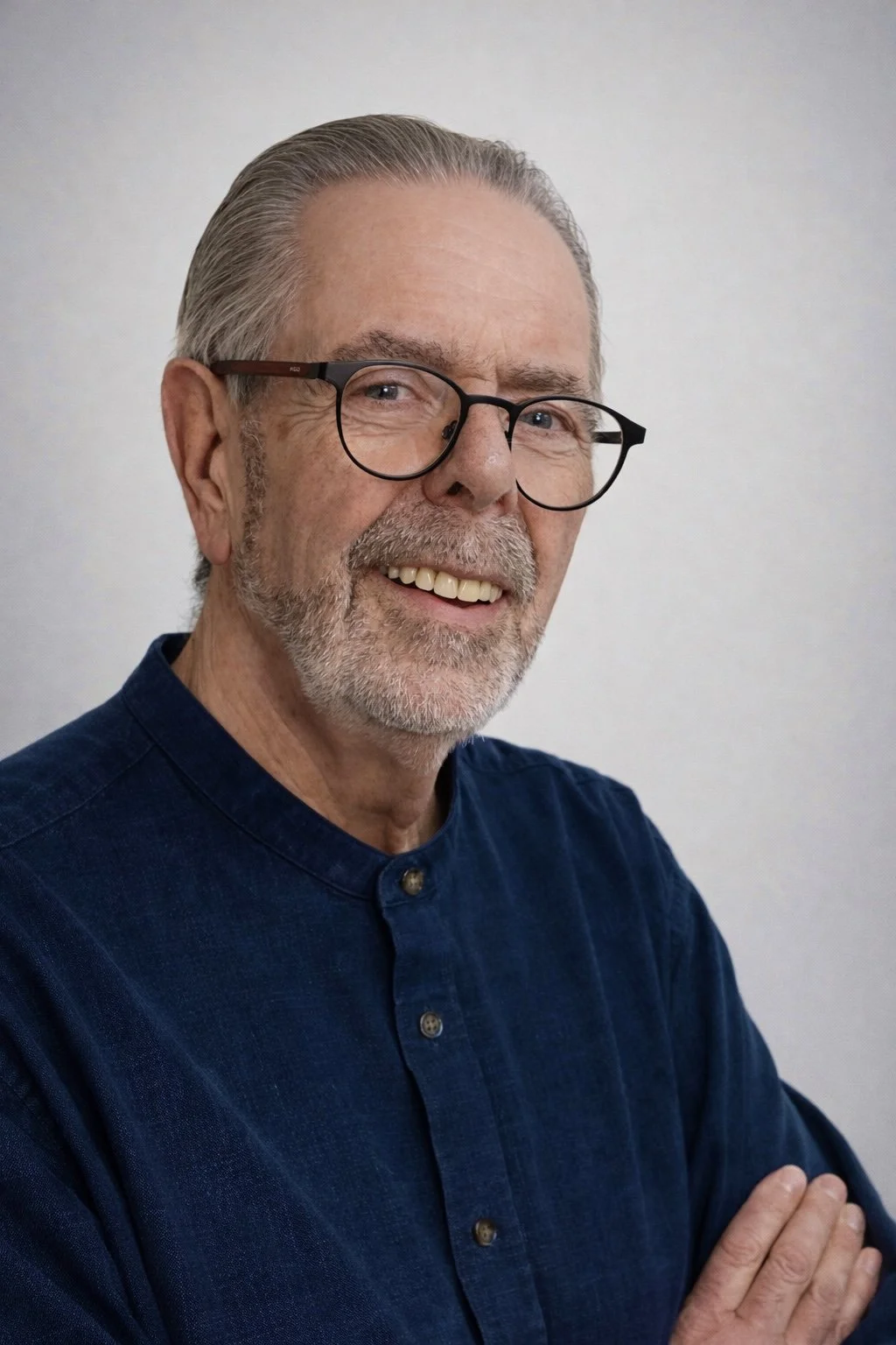 A smiling older man with gray hair, beard, and glasses, wearing a dark blue shirt, standing against a plain white wall.