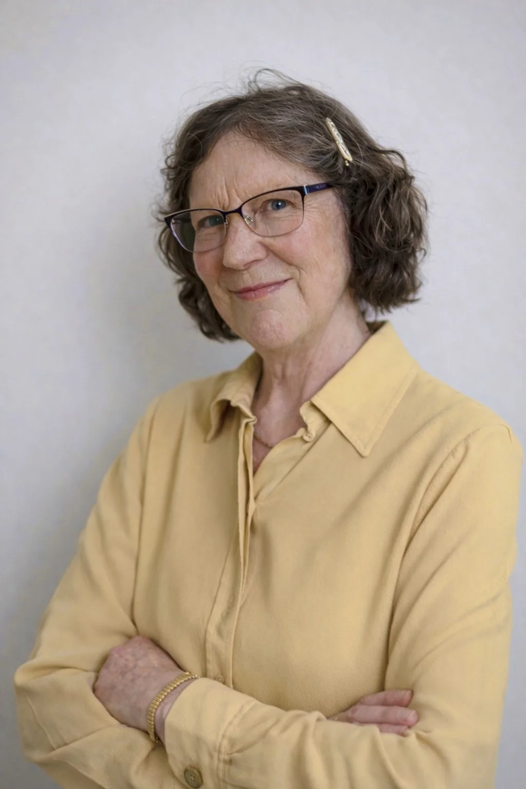 An elderly woman with curly gray hair and glasses, wearing a yellow shirt, standing with arms crossed against a plain light-colored background.
