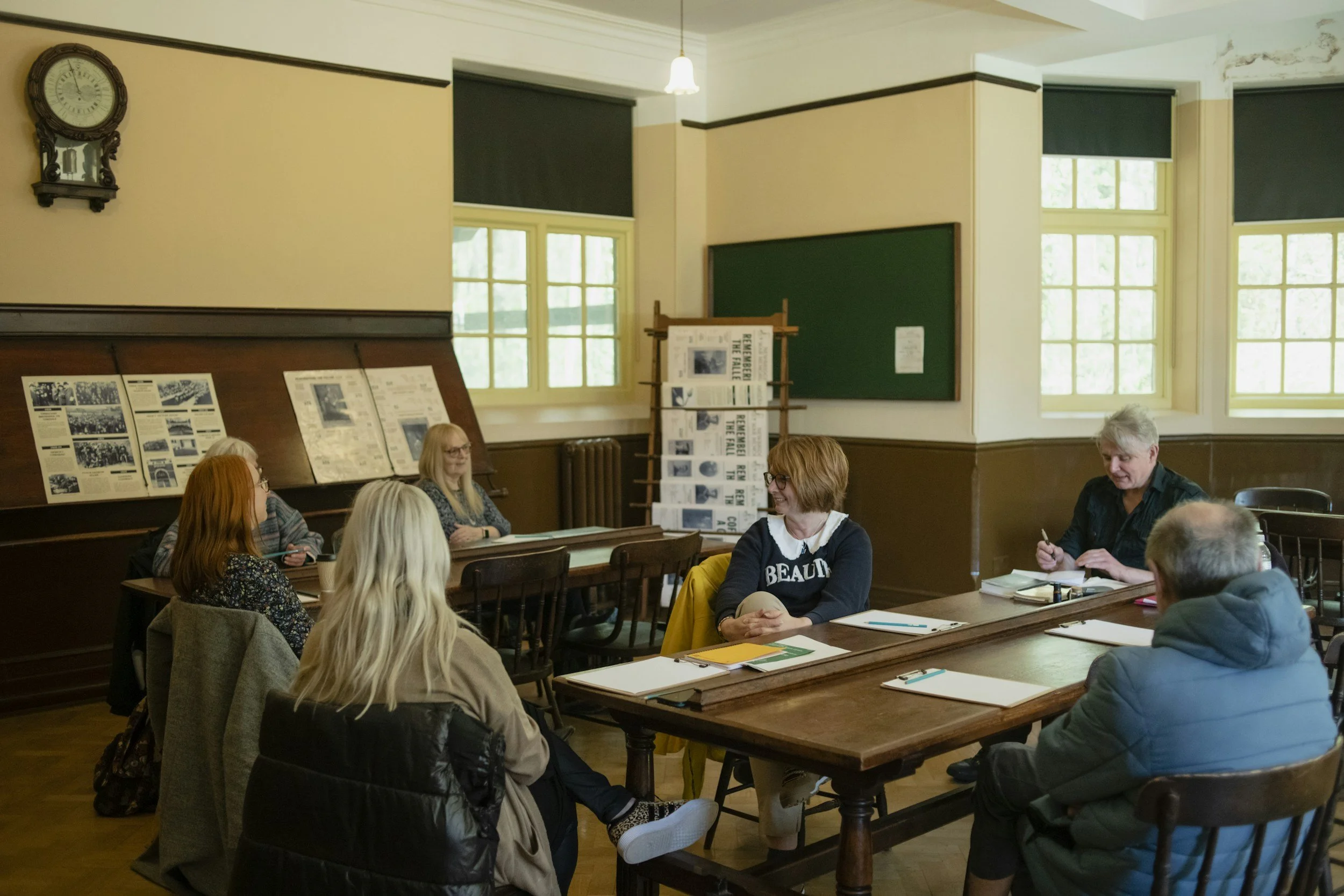 People sitting around tables in a classroom or meeting room with informational posters on a display board, large windows, and a clock on the wall.