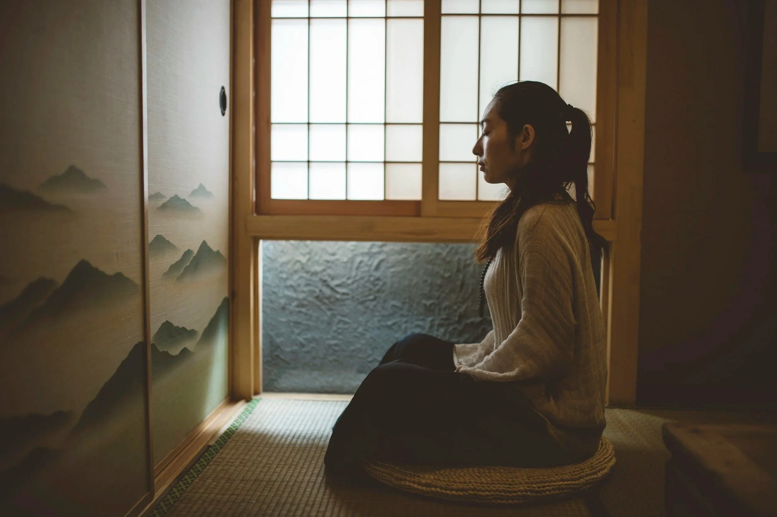 A woman dressed in traditional Japanese attire kneeling on a woven mat inside a room with shoji screen doors, looking at a mountain painting on sliding wooden doors.