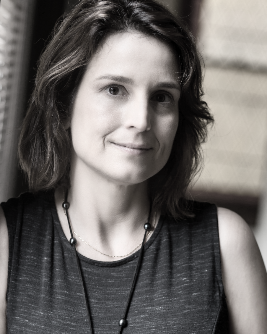 Black and white photo of a woman with shoulder-length hair, wearing a sleeveless top and a necklace, smiling slightly, in an indoor setting.