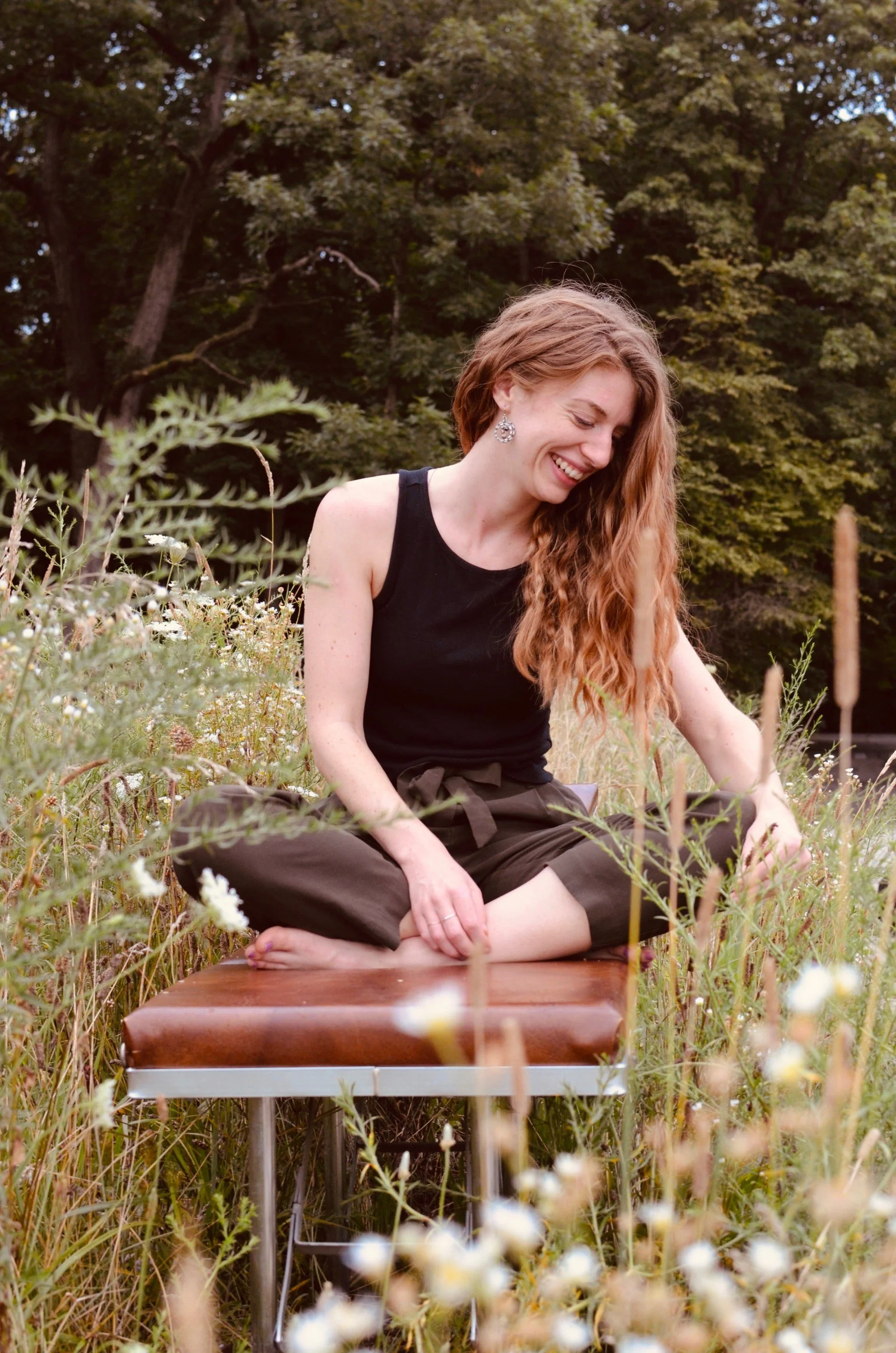 A woman with long, curly red hair and earrings, wearing a black tank top and dark pants, is sitting cross-legged on a brown leather chair in a grassy field with wildflowers, smiling while reaching out to touch the plants. Dense trees are in the background.