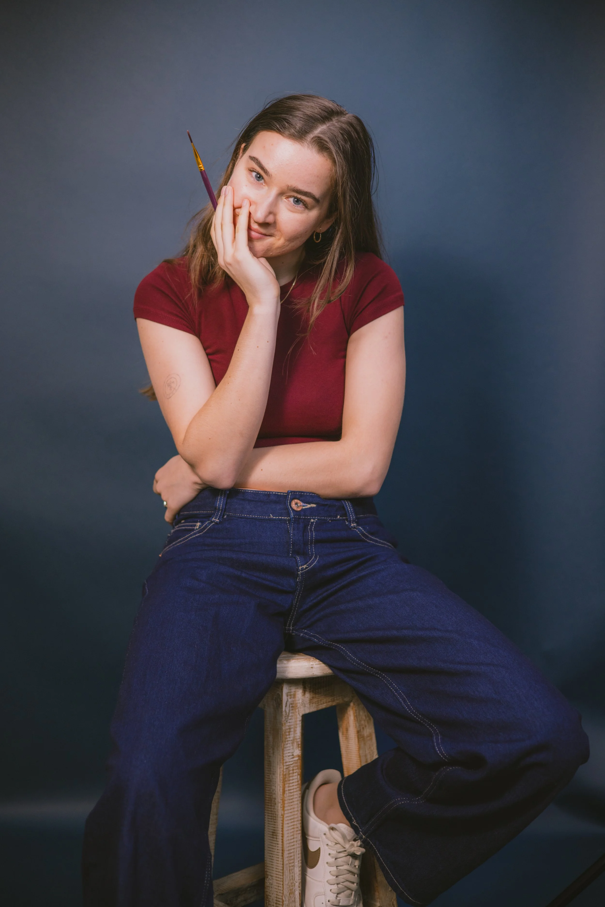 A young woman with brown hair, wearing a red t-shirt, blue jeans, and white sneakers, sitting on a wooden stool against a dark blue background. She has a questioning or playful expression, with one hand resting on her cheek and holding a paintbrush.
