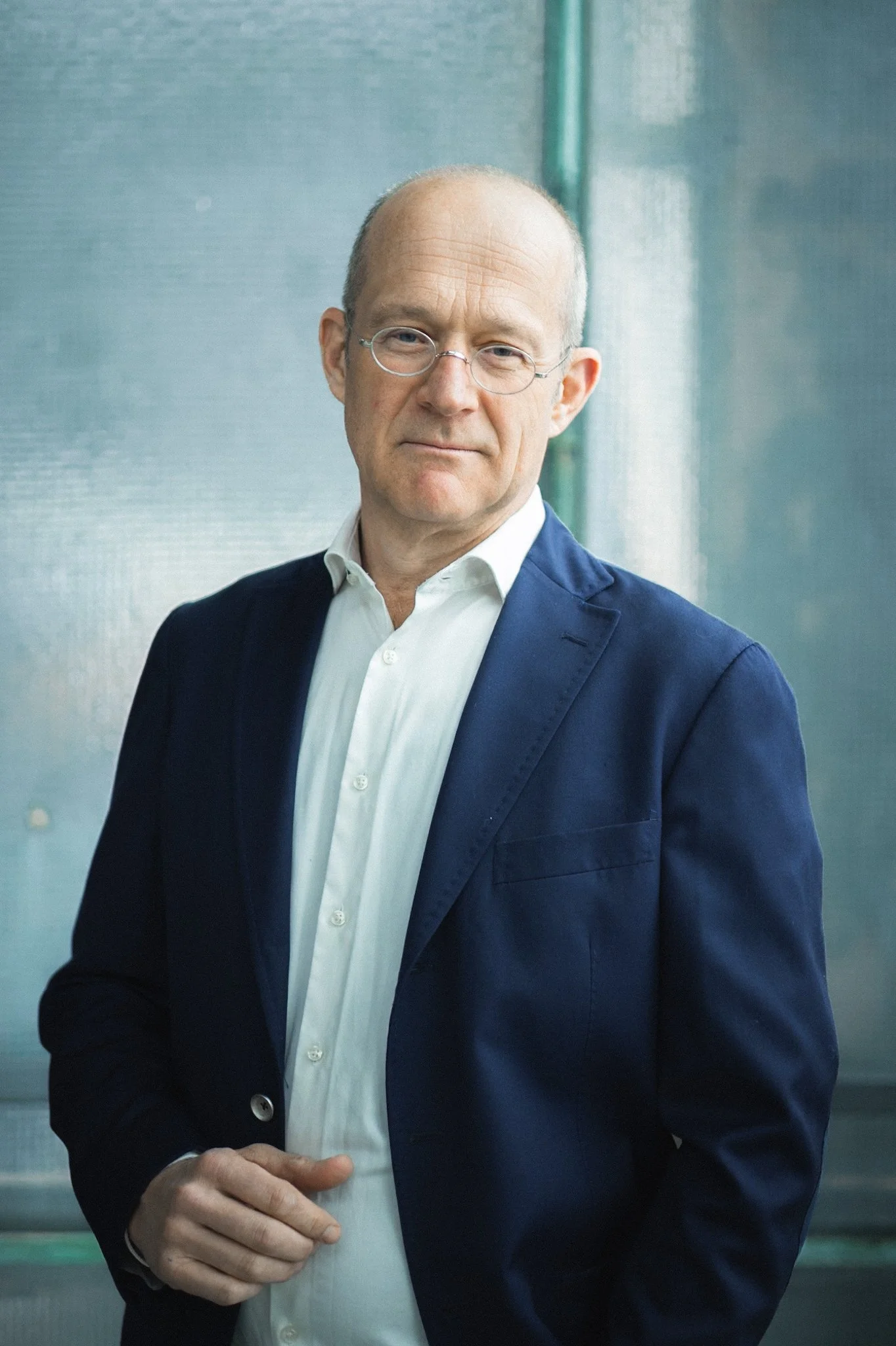 A middle-aged man with glasses wearing a white shirt and navy blazer, standing in front of a blurred background with greenish tones.