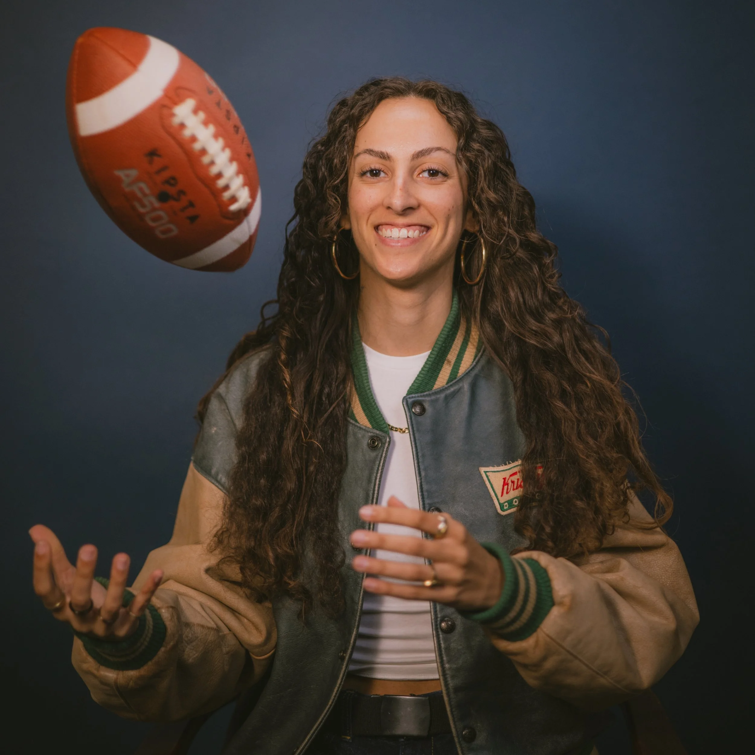 A woman with curly hair smiling, holding a smartphone in her left hand, wearing a varsity jacket with patches, and gesturing with her right hand against a dark background.