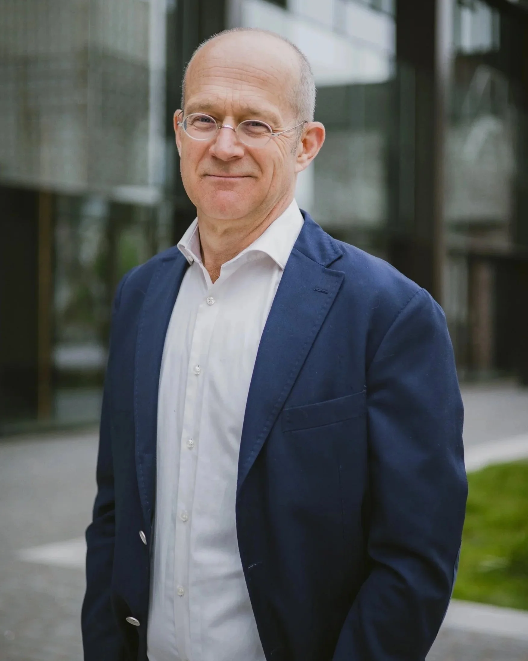 A middle-aged man with glasses, wearing a white shirt and a dark blue blazer, standing outdoors in front of a modern glass building.