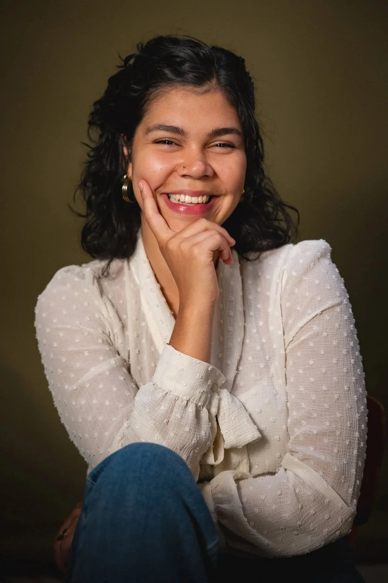 A woman with dark curly hair smiling and resting her chin on her hand, wearing a white textured blouse with puffed sleeves, seated against a plain background.
