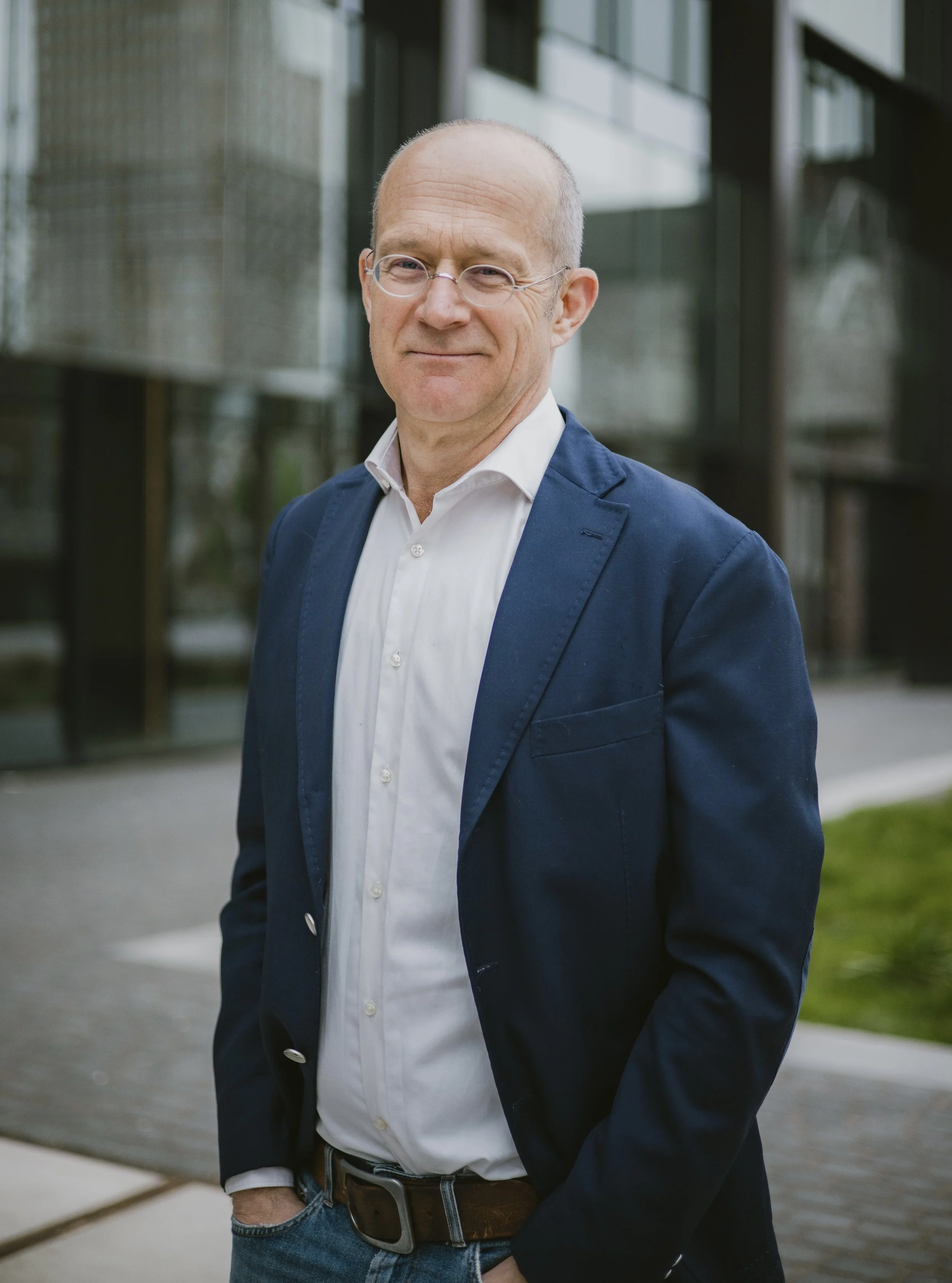 A middle-aged man with glasses wearing a white shirt and navy blazer standing outdoors in front of a modern glass building.