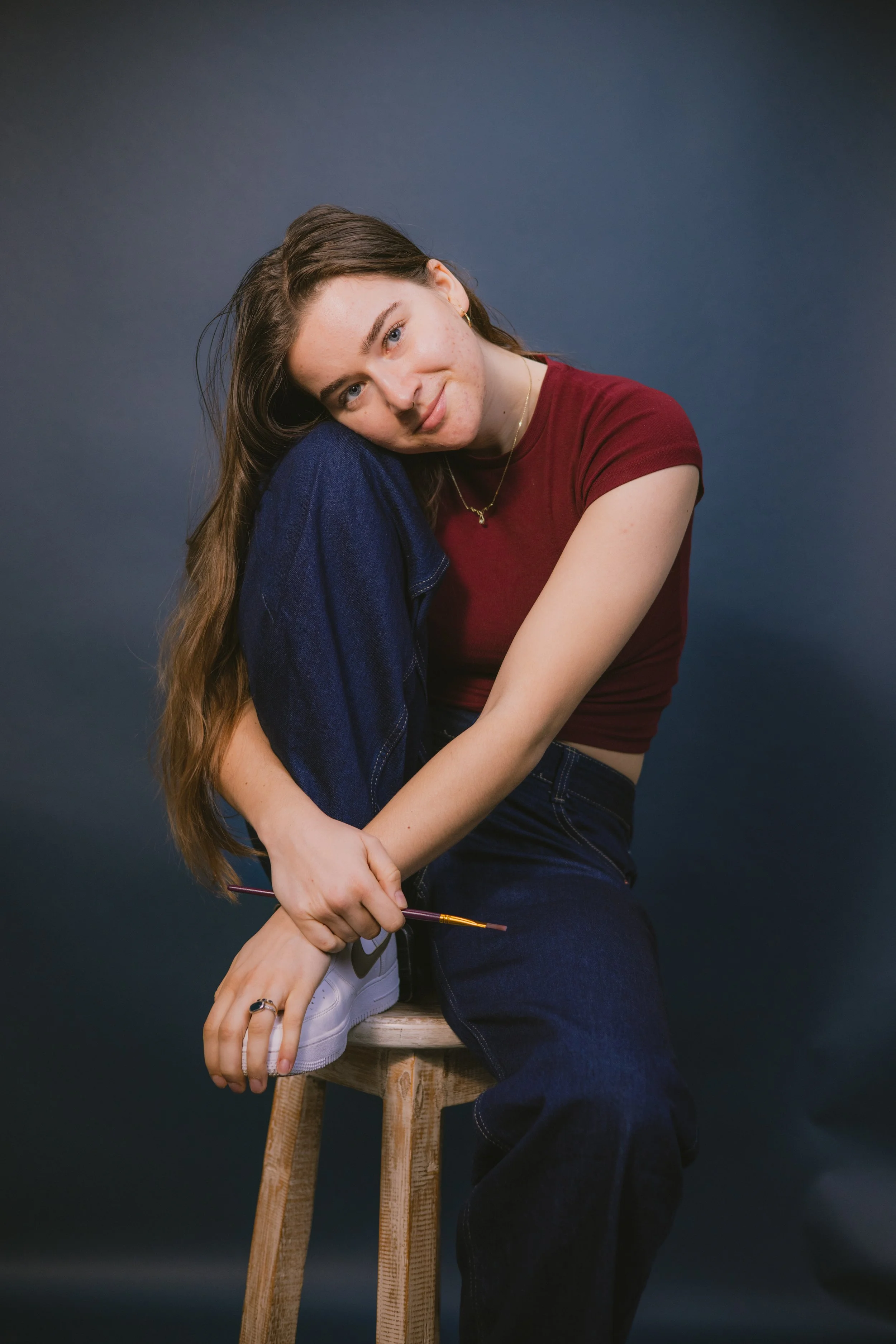 Young woman with long brown hair sitting on a wooden stool, wearing a red crop top, dark jeans, and white sneakers, with a dark background.