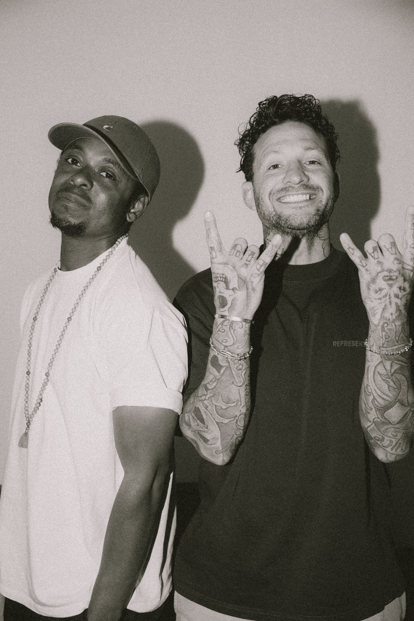 Two men standing side by side, the man on the left wearing a baseball cap, a white t-shirt, and a beaded necklace, with a serious expression. The man on the right has short, curly hair, tattoos on his arms and hands, and is smiling broadly while making a peace sign with both hands.