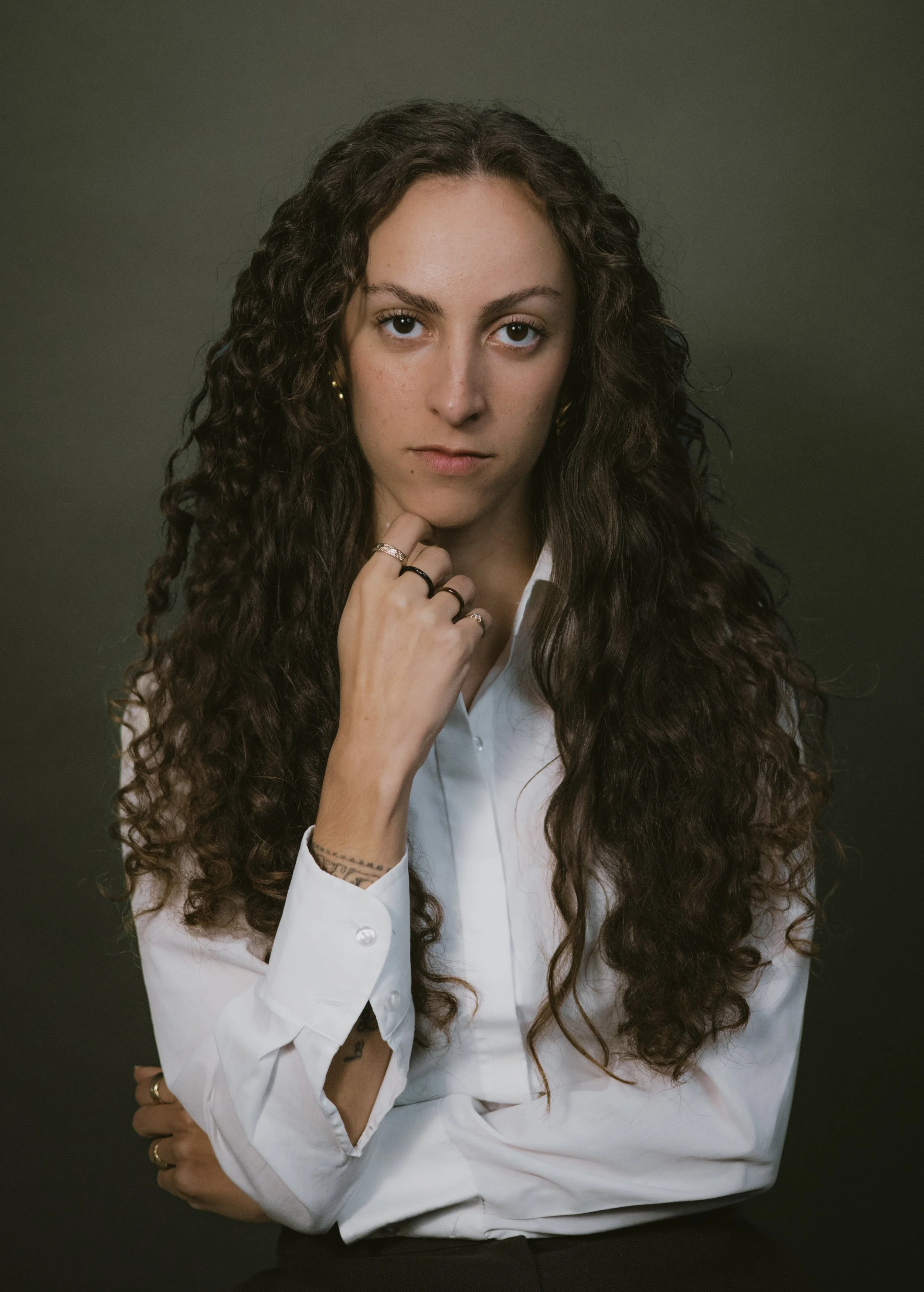 A confident woman with long curly dark hair wearing a white shirt against a dark background.