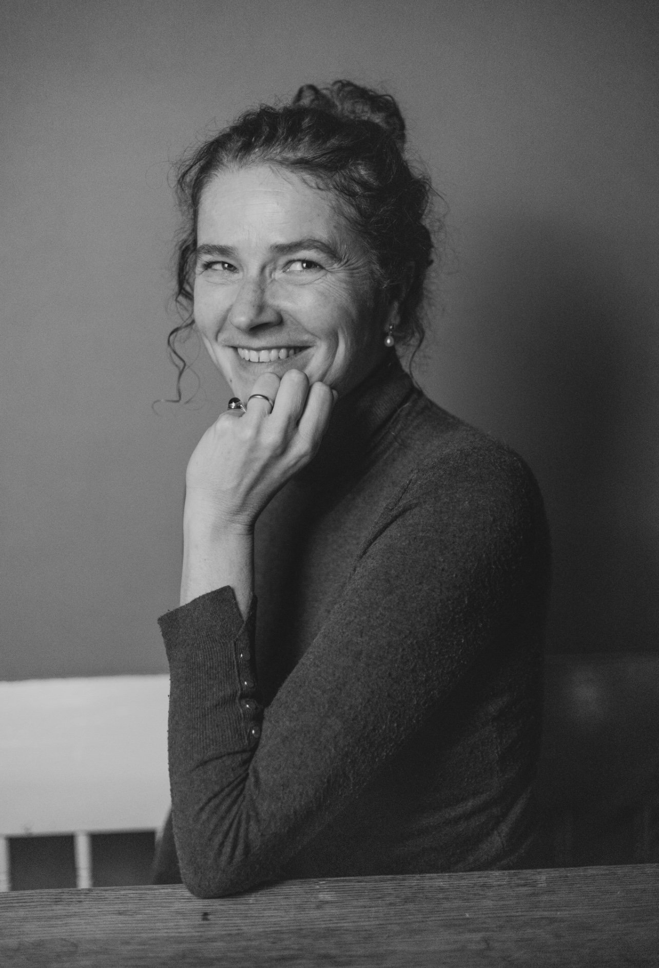 Black and white portrait of a woman smiling, resting her chin on her hand, wearing a long-sleeve top, with earrings and rings.