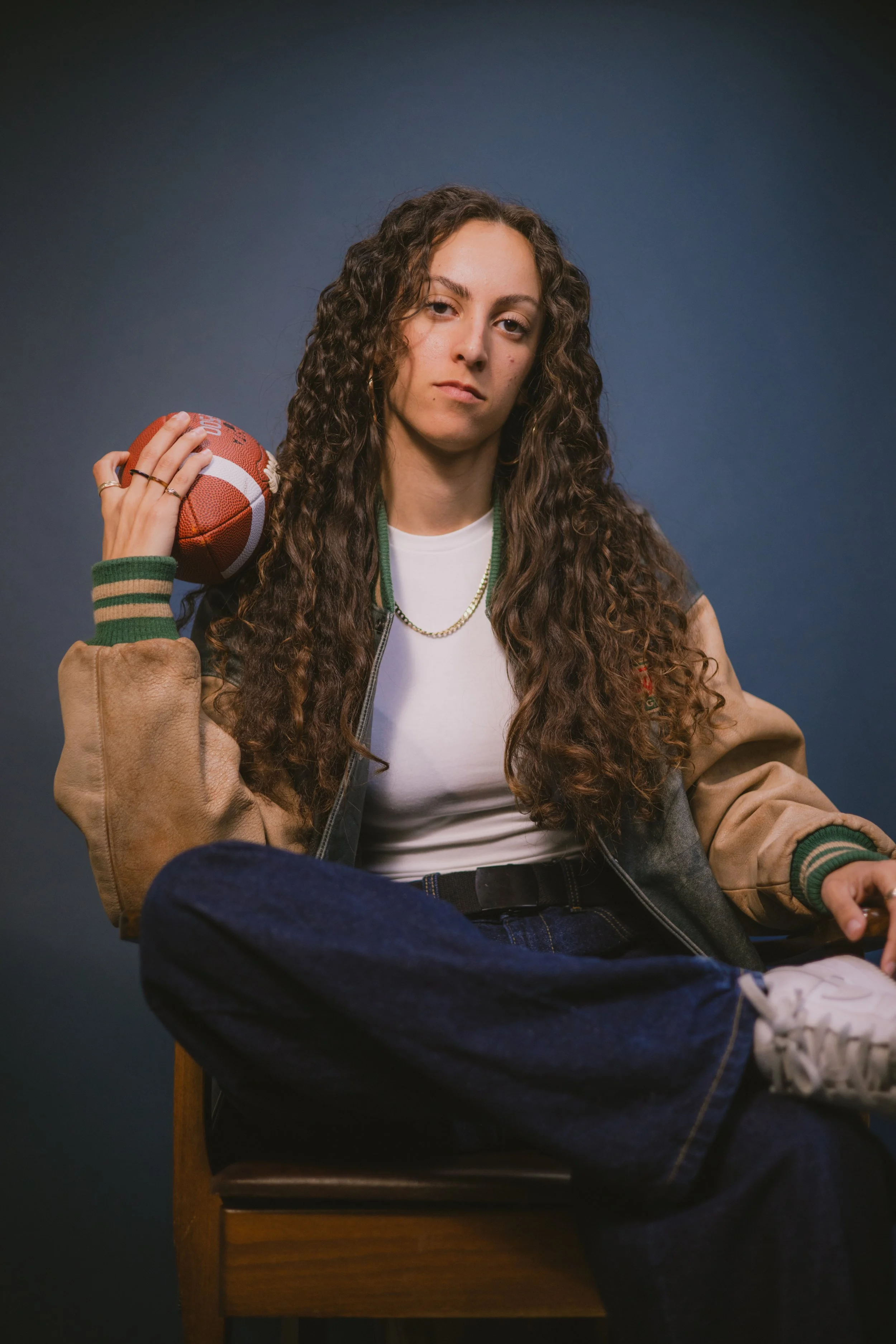 A young woman with long curly hair, wearing a varsity jacket, white t-shirt, and jeans, seated with one leg crossed, holding a football over her shoulder, against a dark gray background.