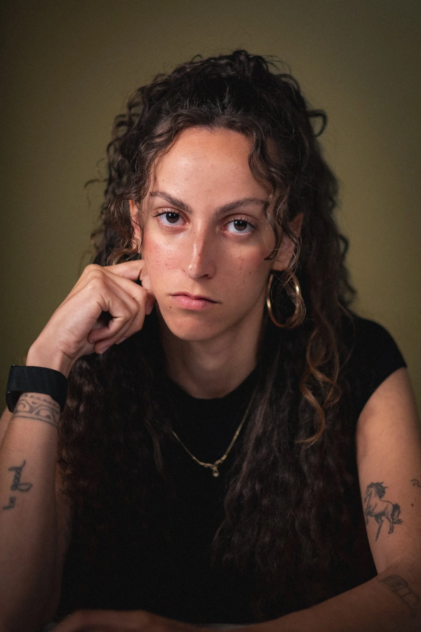 A woman with curly brown hair and tattoos on her arms resting her chin on her hand, wearing a black top and gold jewelry, looking directly into the camera against a plain background.