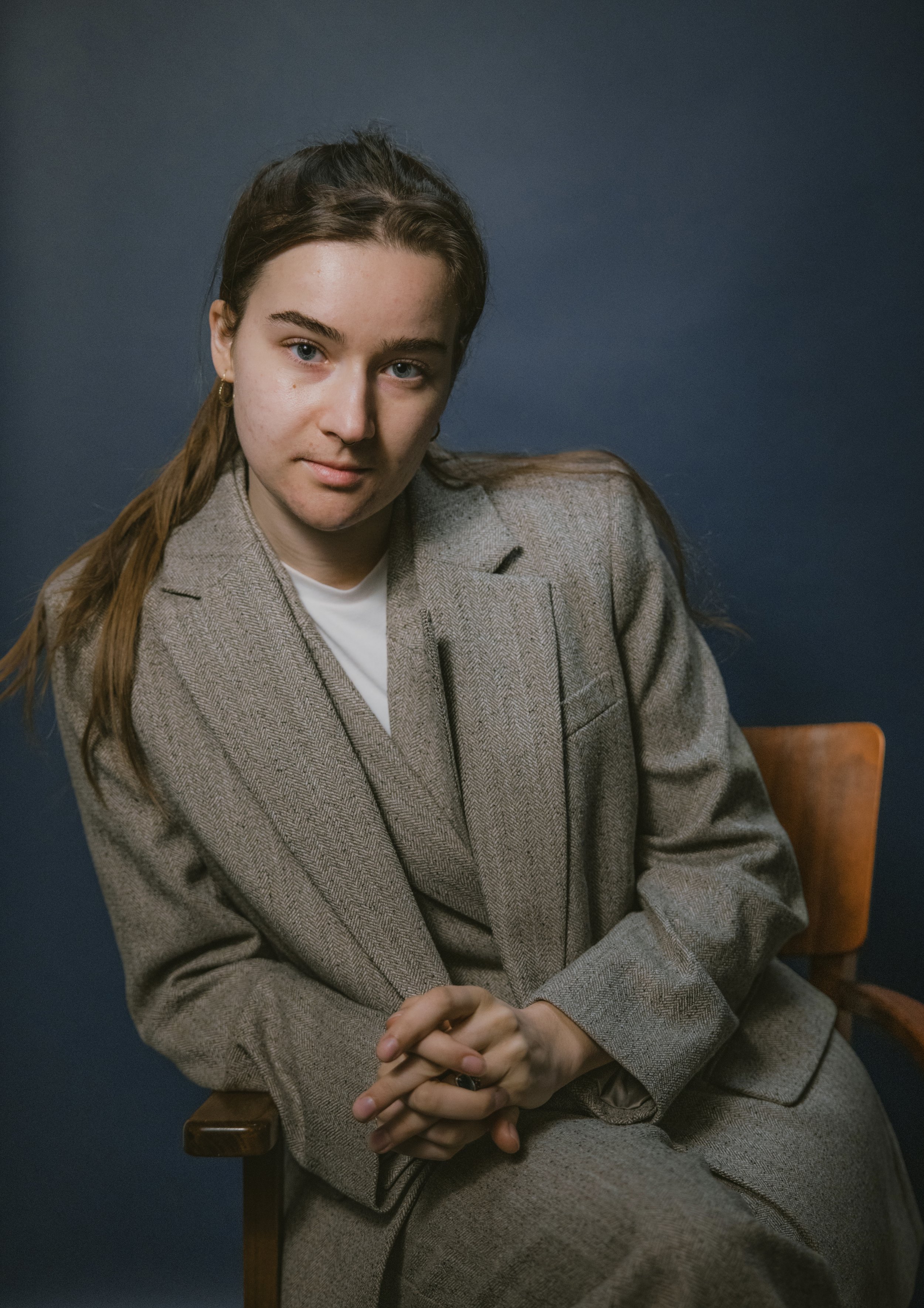A young woman with long brown hair, blue eyes, and light skin wearing a gray blazer, sitting with her hands clasped, against a dark blue background.