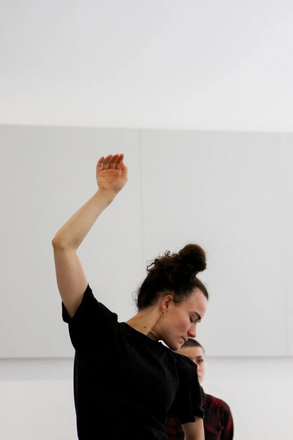 A woman with curly hair in a bun performs a dance move with her arm raised in a studio with white walls.