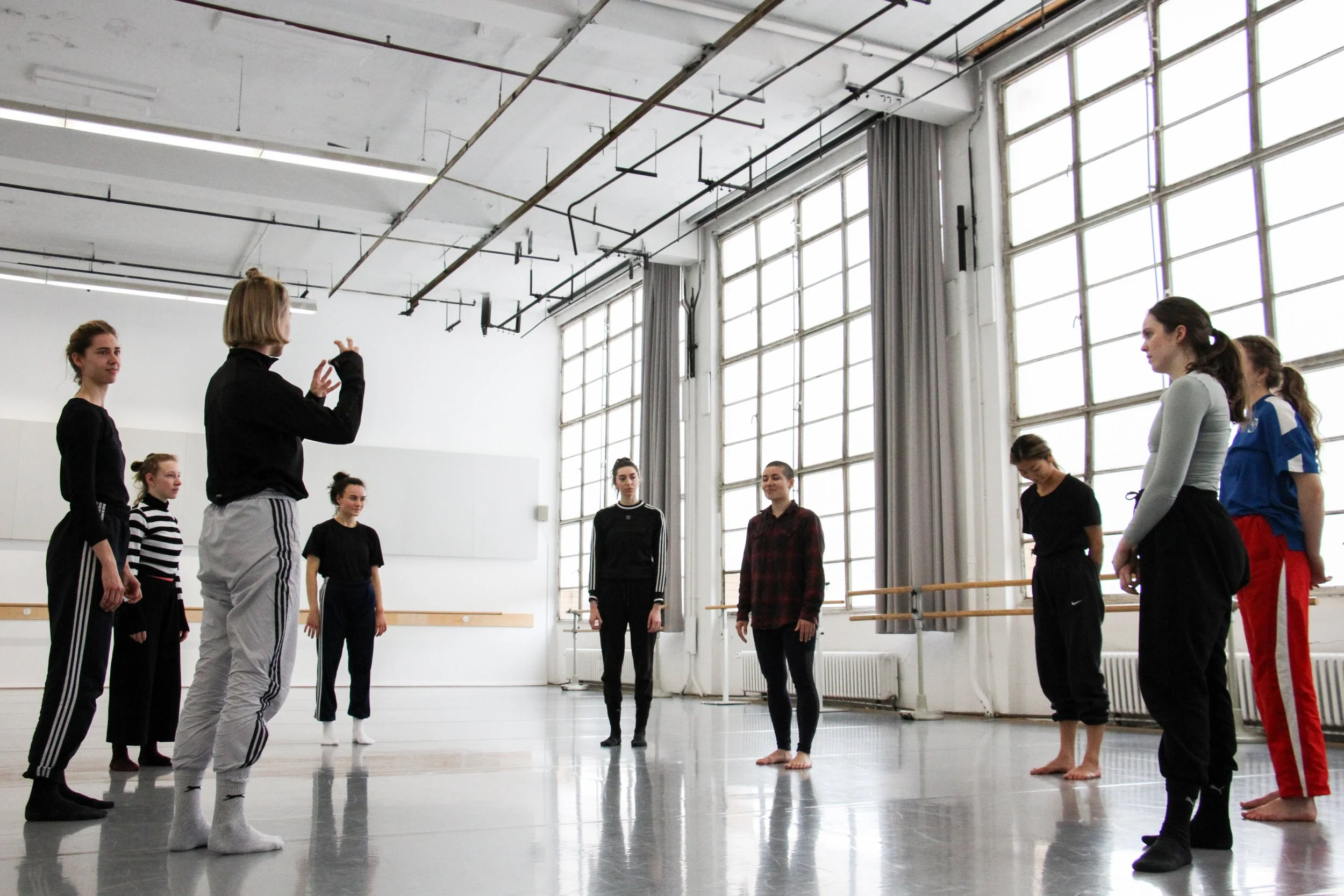 A group of dancers standing in a dance studio with large windows, listening to an instructor who appears to be giving directions.
