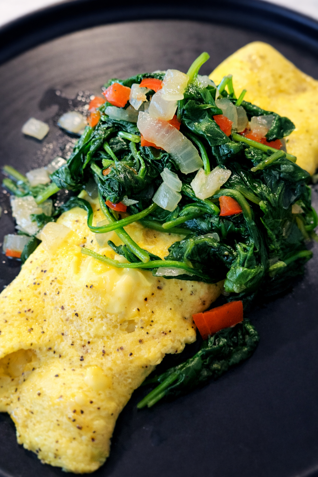 Close-up of a yellow omelette topped with sautéed spinach and diced vegetables, served on a black plate.