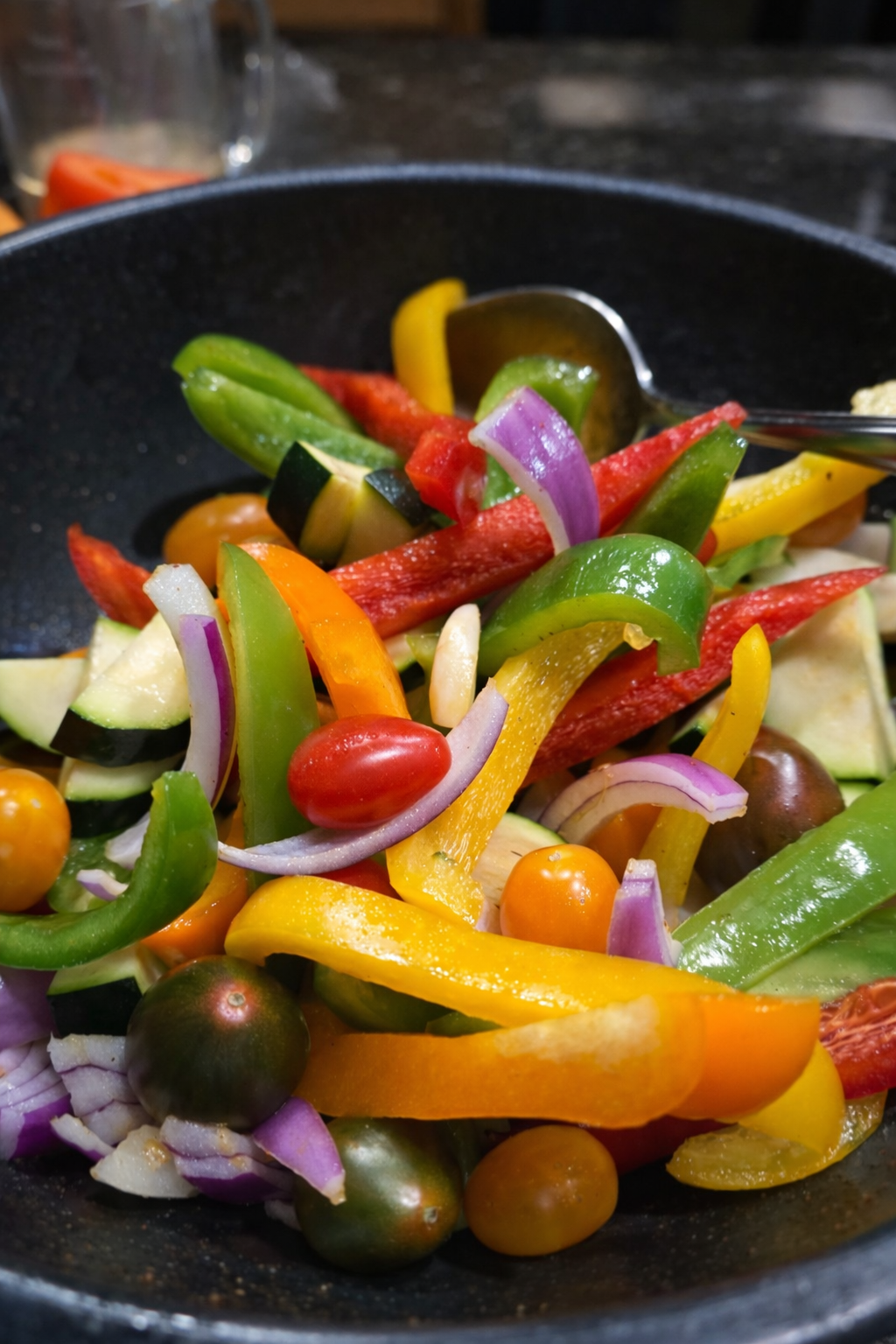 Colorful sliced vegetables including red, green, yellow, and purple peppers, cherry tomatoes, and red onions in a frying pan.