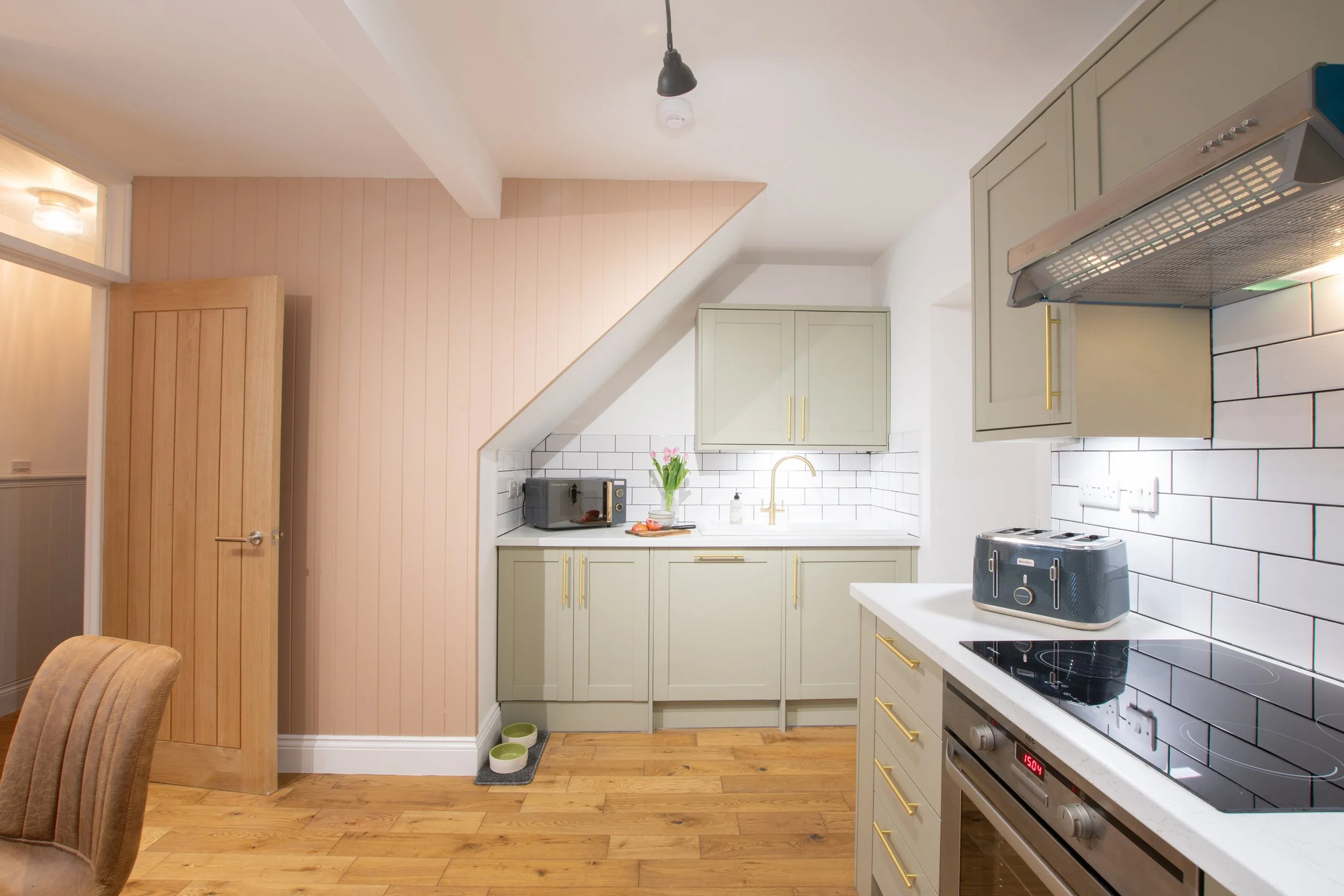 A modern kitchen with green cabinets, white subway tile backsplash, black stove, and wooden floor. There are three pet bowls on a small rug, and a microwave, toaster, and vase with pink tulips on the counter.