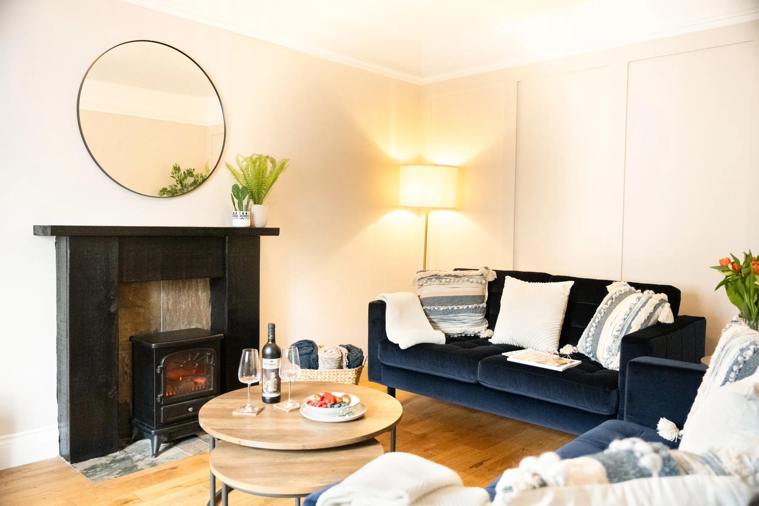A cozy living room with a black sofa with white and blue cushions, a wooden coffee table with a bowl of berries and two wine glasses, a fireplace with a black mantle, a potted fern plant, a mirror on the wall, a standing lamp, and a basket with blank
