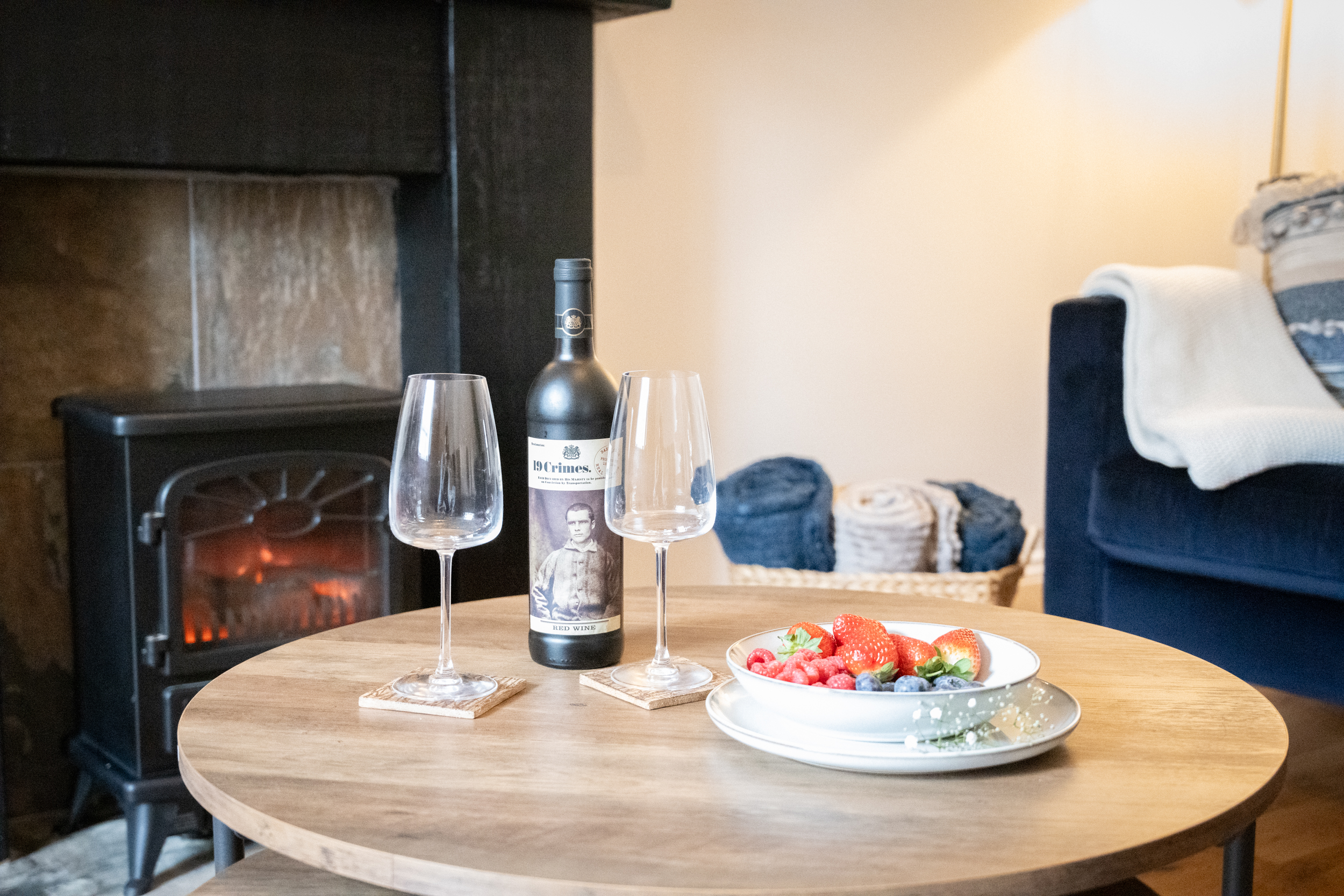 A round wooden table set with a bottle of red wine, two empty wine glasses, and a bowl of fresh strawberries and other berries. In the background, a cozy living room features a black fireplace with a warm fire, a basket of blankets, and a couch with 