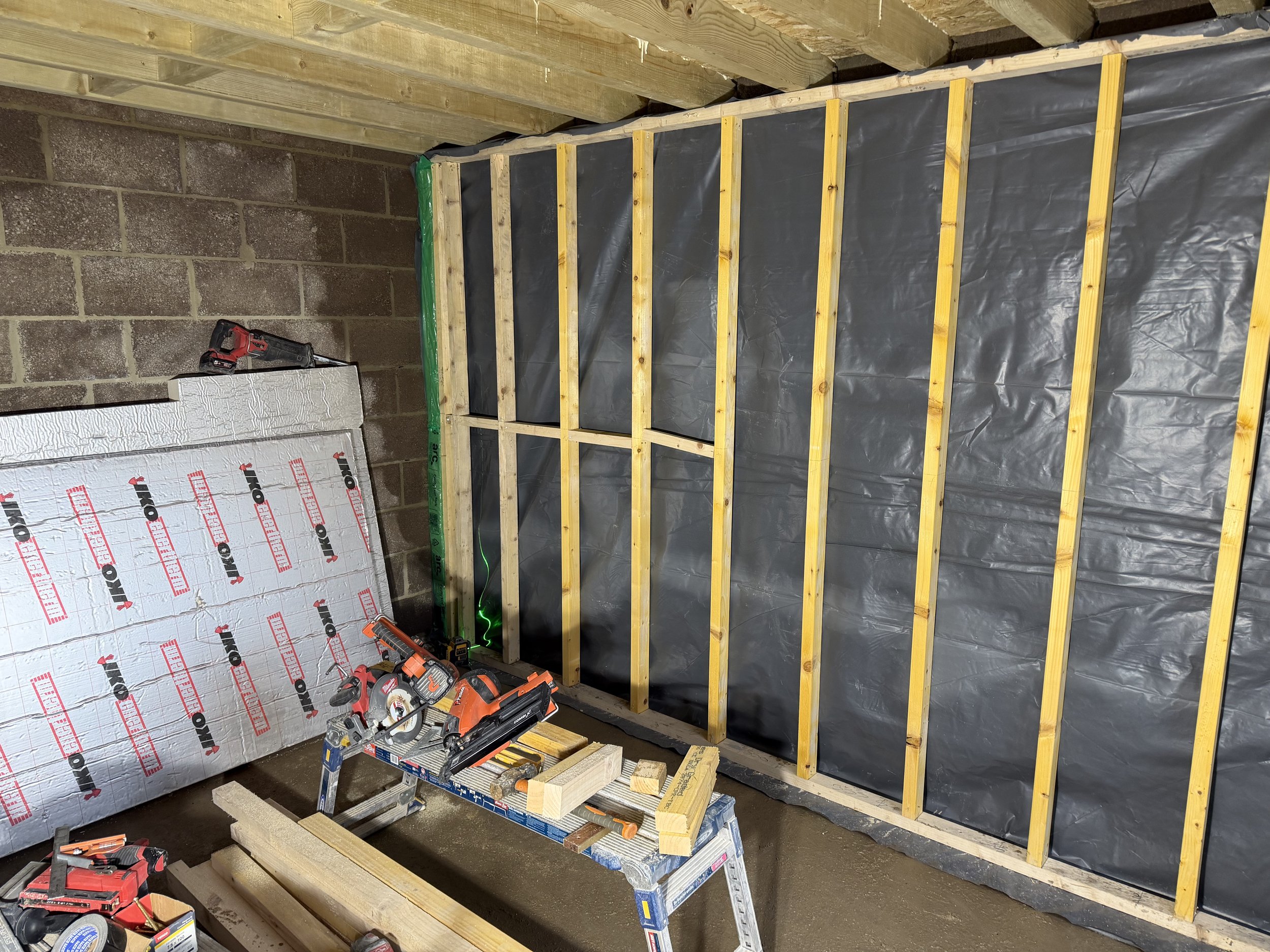 An indoor construction site showing wooden framing for a wall with black sheathing. There are tools and materials on a workbench and the floor, including power drills, a saw, and insulation panels.