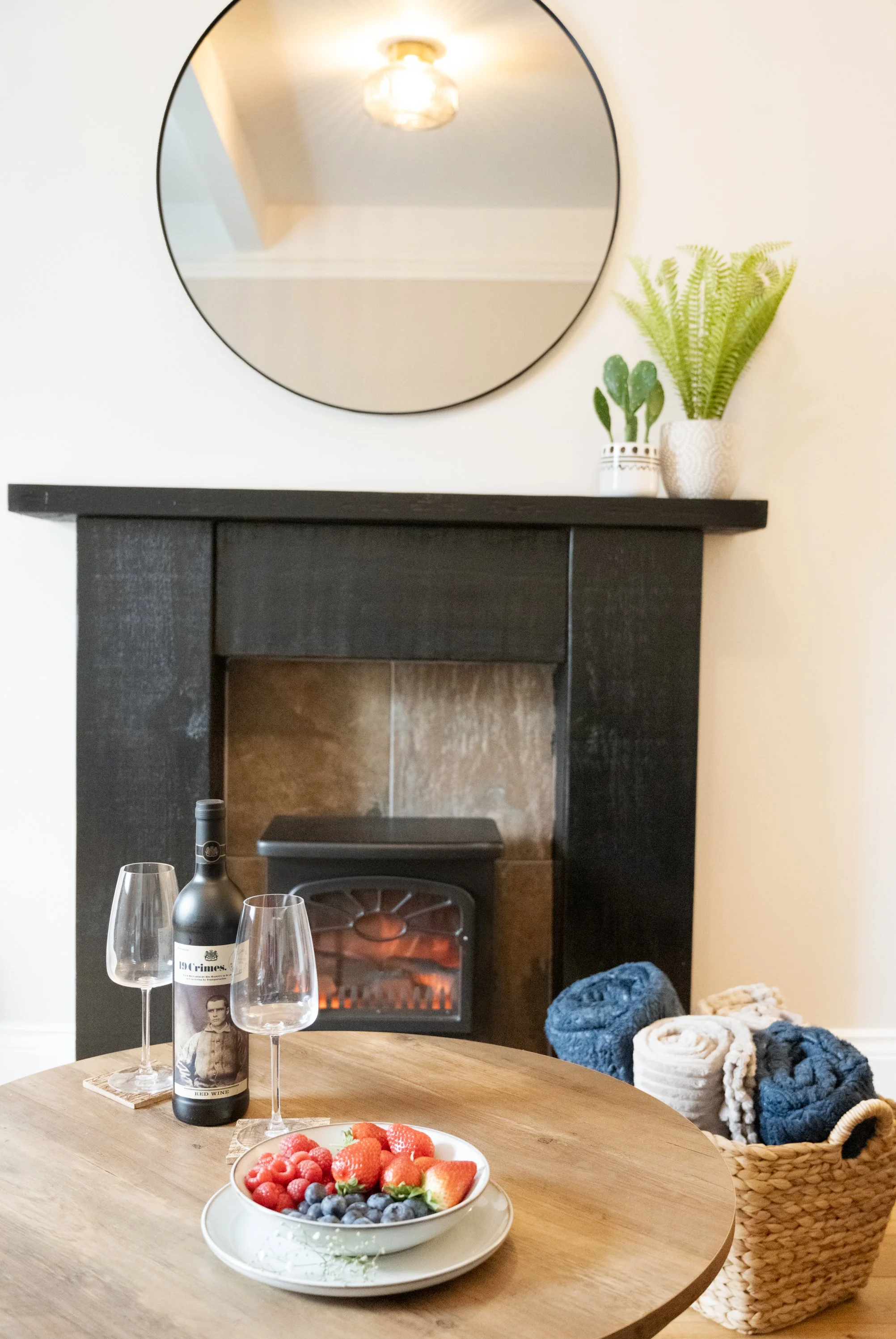 A round wooden table with a bowl of strawberries, raspberries, and blueberries, two wine glasses, and a bottle of red wine. In the background, there is a fireplace, a mirror on the wall, and potted plants on the mantelpiece. A basket with rolled-up b