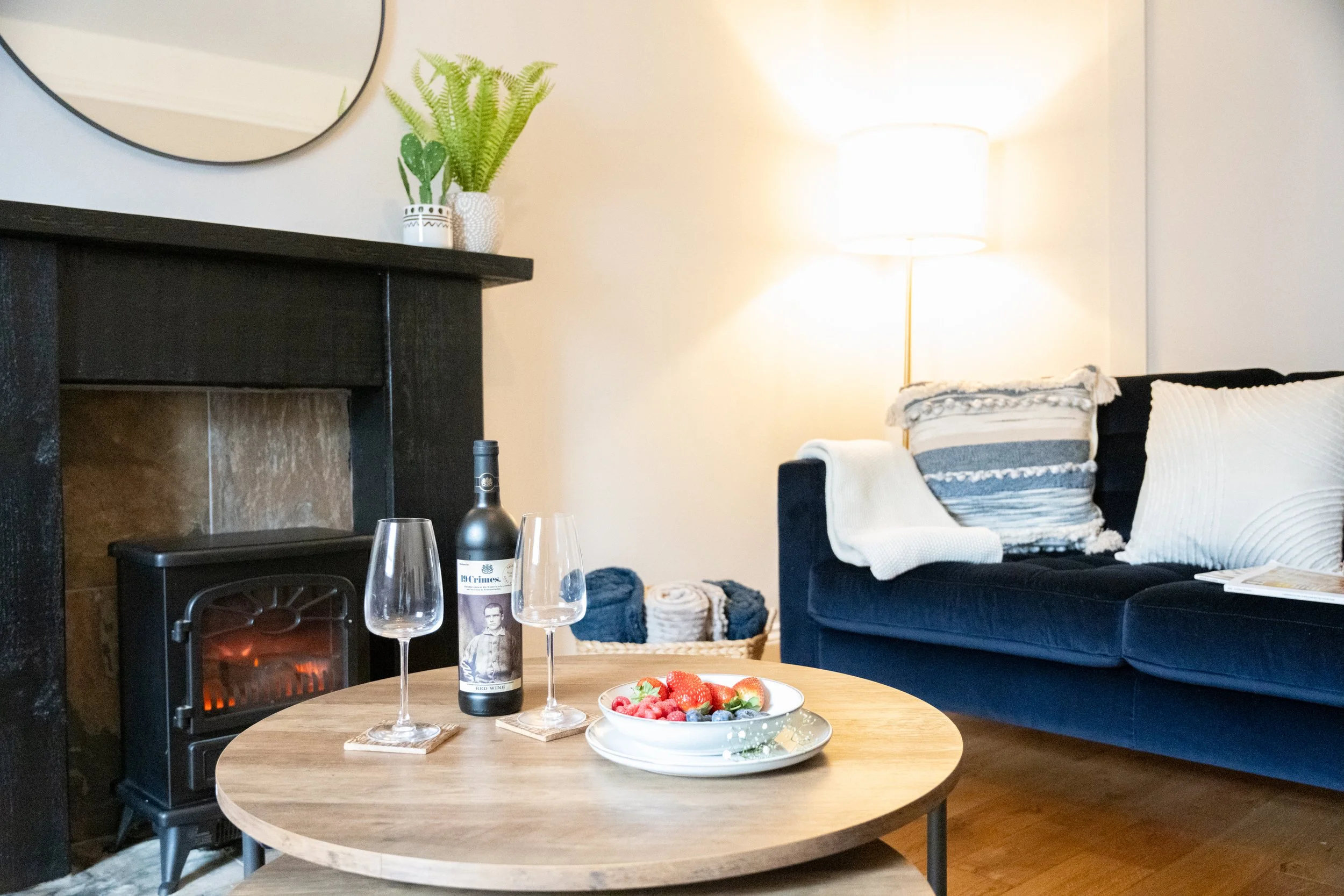 Living room with a round wooden coffee table holding a bowl of mixed berries, two wine glasses, and a wine bottle. There is a fireplace with a fire burning, a black sofa with white pillows and a blanket, a side table with a lamp, and decorative plant
