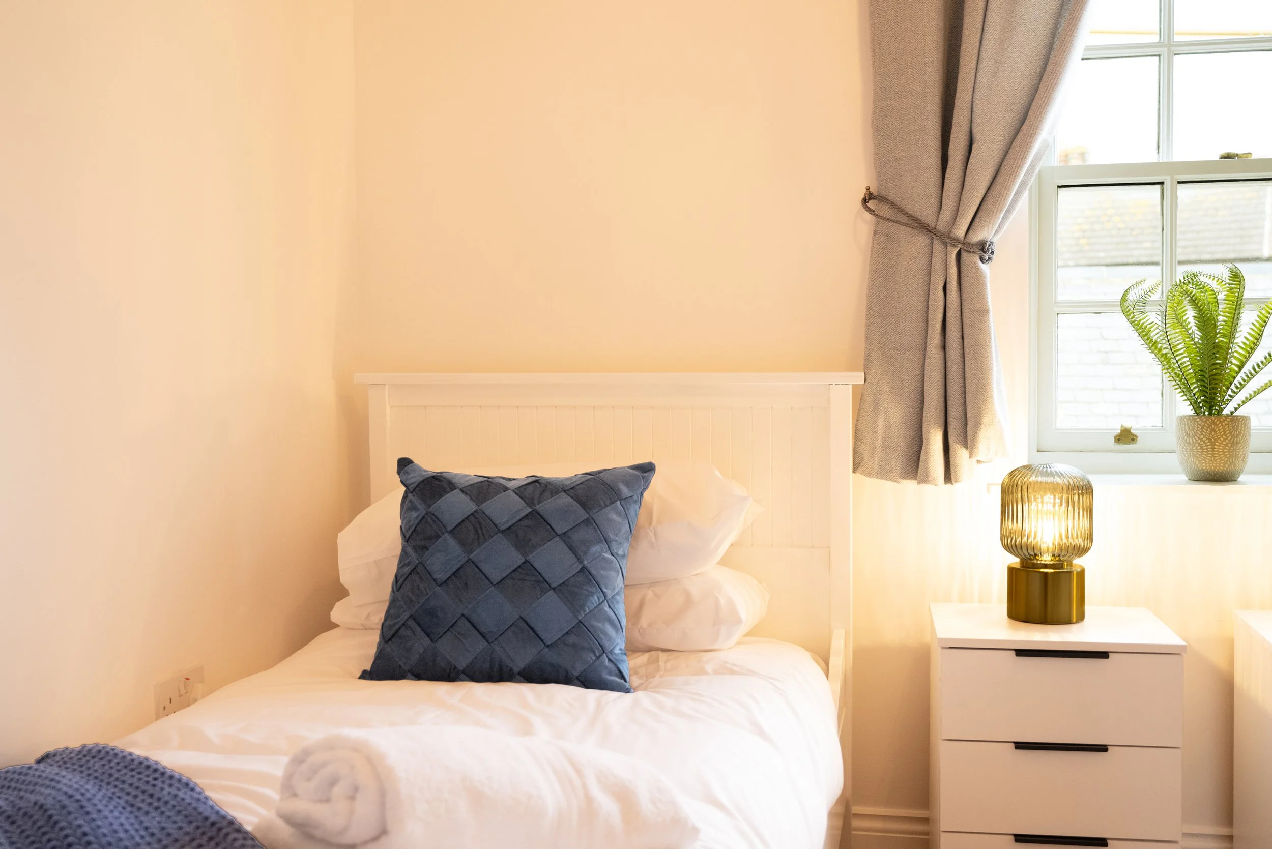 A cozy bedroom with a white bed, topped with white and dark blue pillows, next to a white nightstand with a gold lamp. A window with gray curtains and a potted fern on the windowsill add natural light.