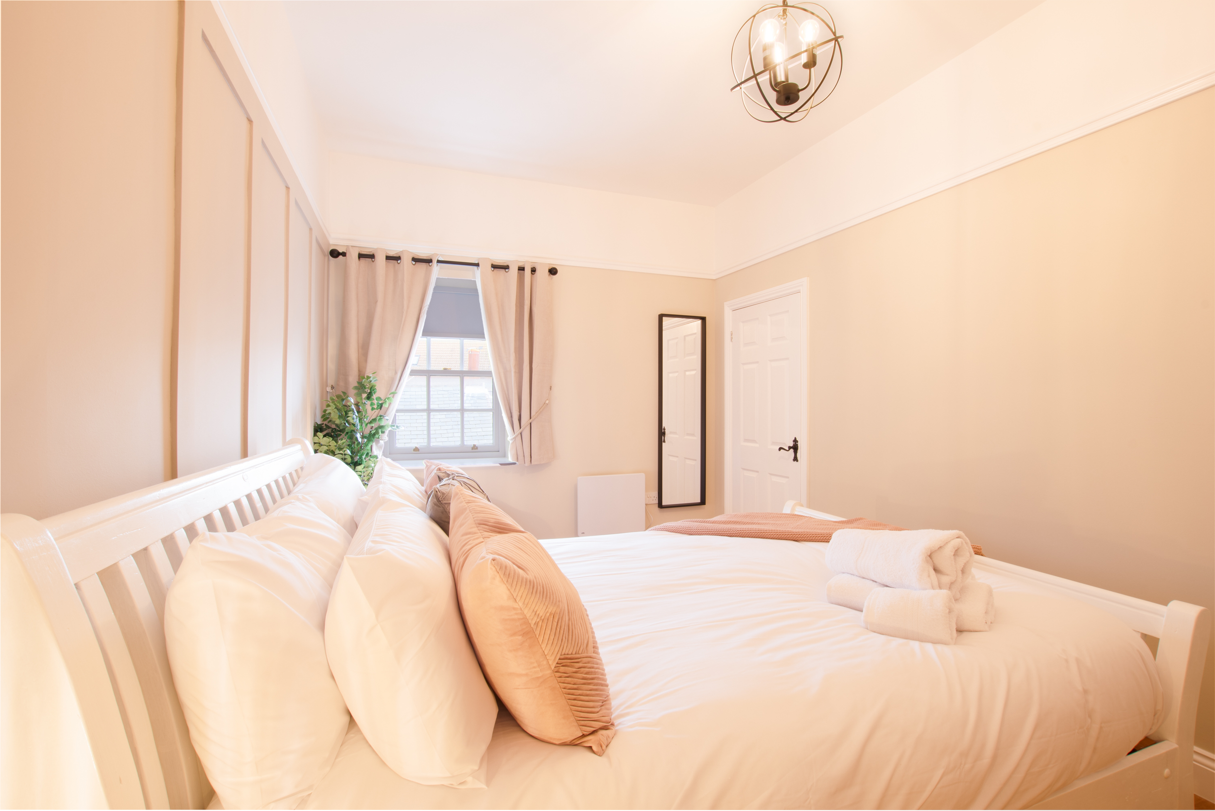 A cozy bedroom with a neatly made bed, cream-colored walls, cream curtains, and a window with a view of rooftops. A tall mirror is on the wall, and a potted plant is near the window. The room has a modern ceiling light fixture.