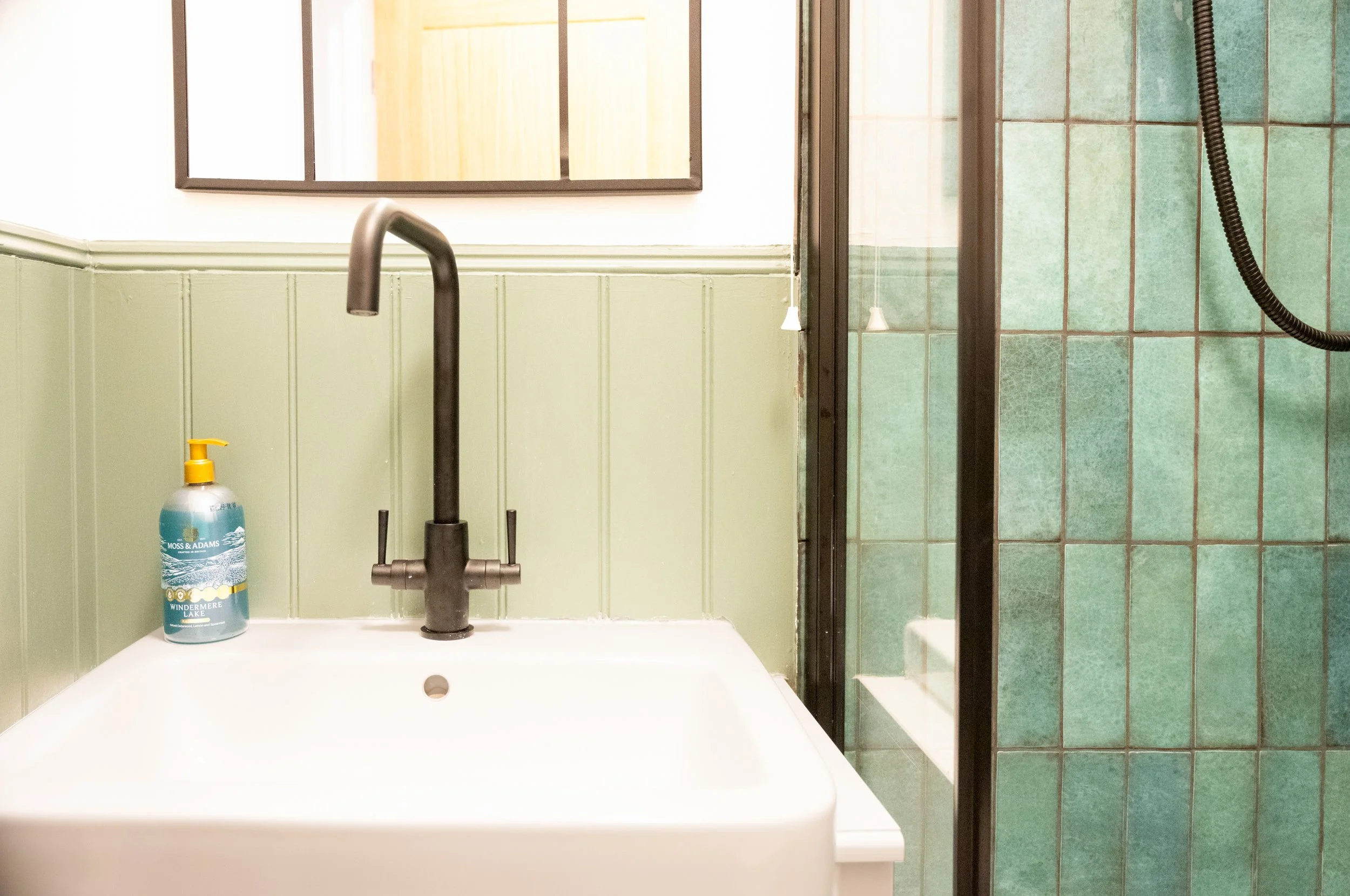 A bathroom sink with a black faucet, a green wall, and a bottle of hand soap on the side of the sink. A mirror and a shower with green tiles and a black shower hose are visible in the background.