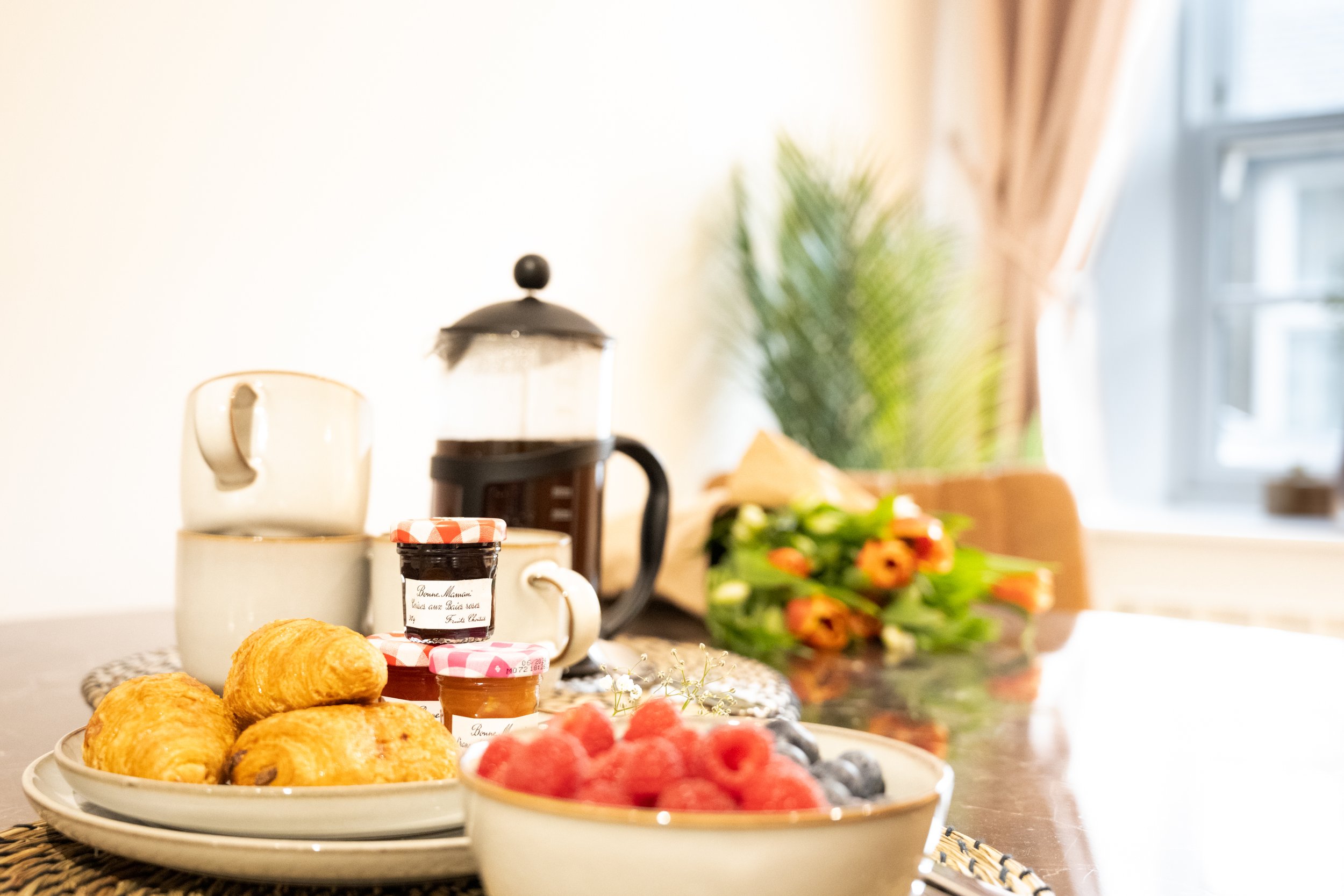 A breakfast table with croissants, bowls of berries, jam jars, a coffee pot, a mug, and a plant near a window with curtains.