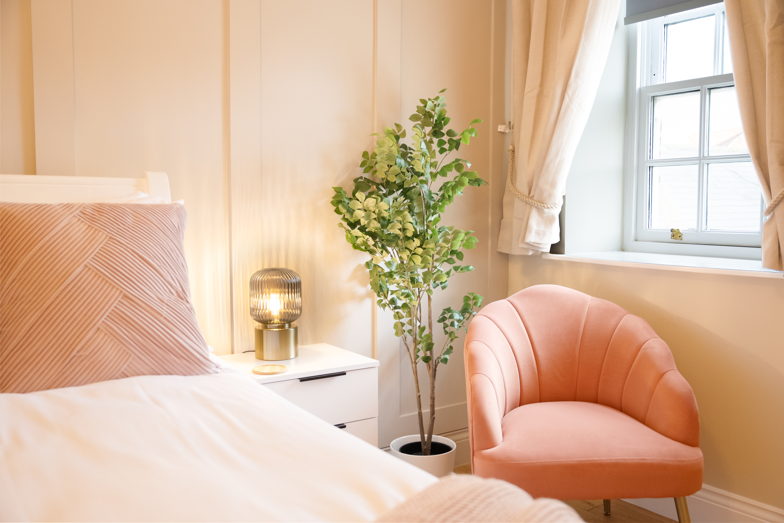 A cozy bedroom corner featuring a white bed with a pink and white striped pillow, a white nightstand, a gold and glass table lamp, a green potted plant, a peach-colored armchair, and a window with cream curtains letting in natural light.