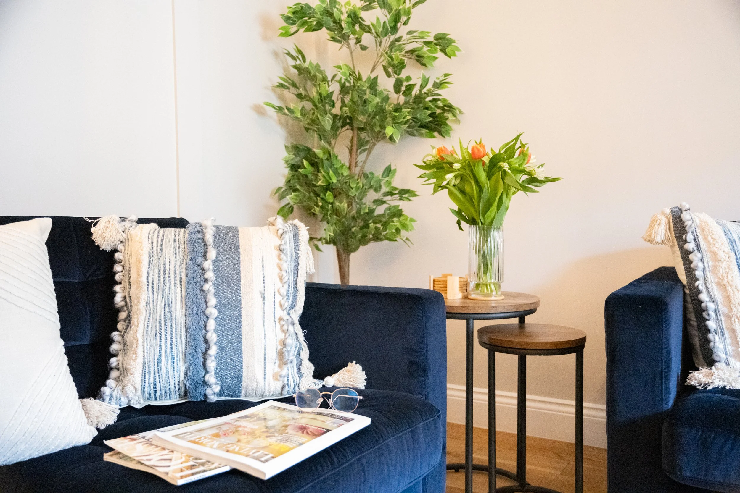 A living room scene featuring a black sofa with decorative pillows, a small round wooden side table with a glass vase of orange and white tulips, a larger green leafy artificial plant in the background, and a pair of glasses resting on a magazine on 