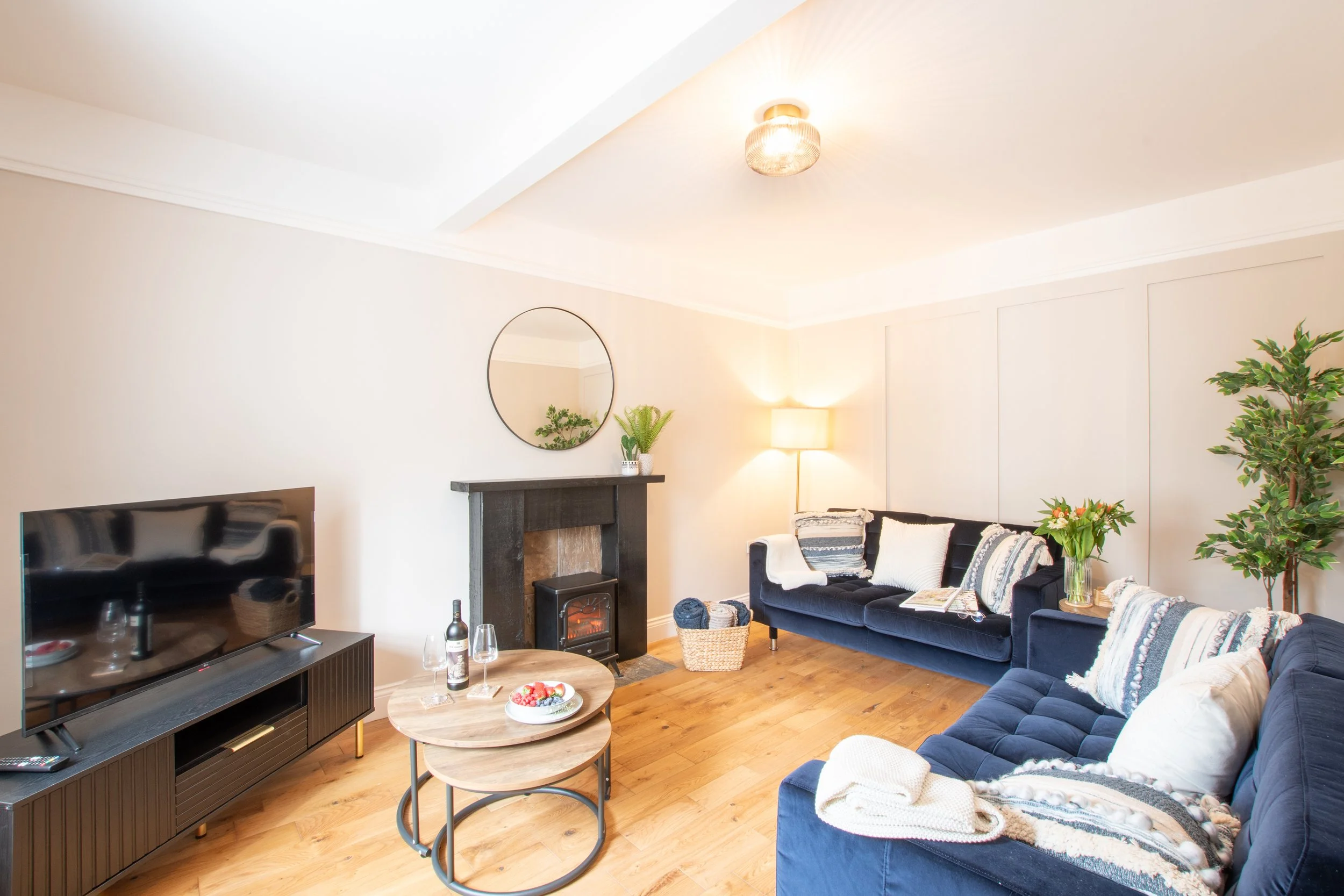 Living room featuring a black sofa set with white and patterned pillows, a round wooden coffee table with a bowl of berries, a TV on a black TV stand, a fireplace, a television remote, a bottle of wine, two wine glasses, a floor lamp, a wall mirror, 