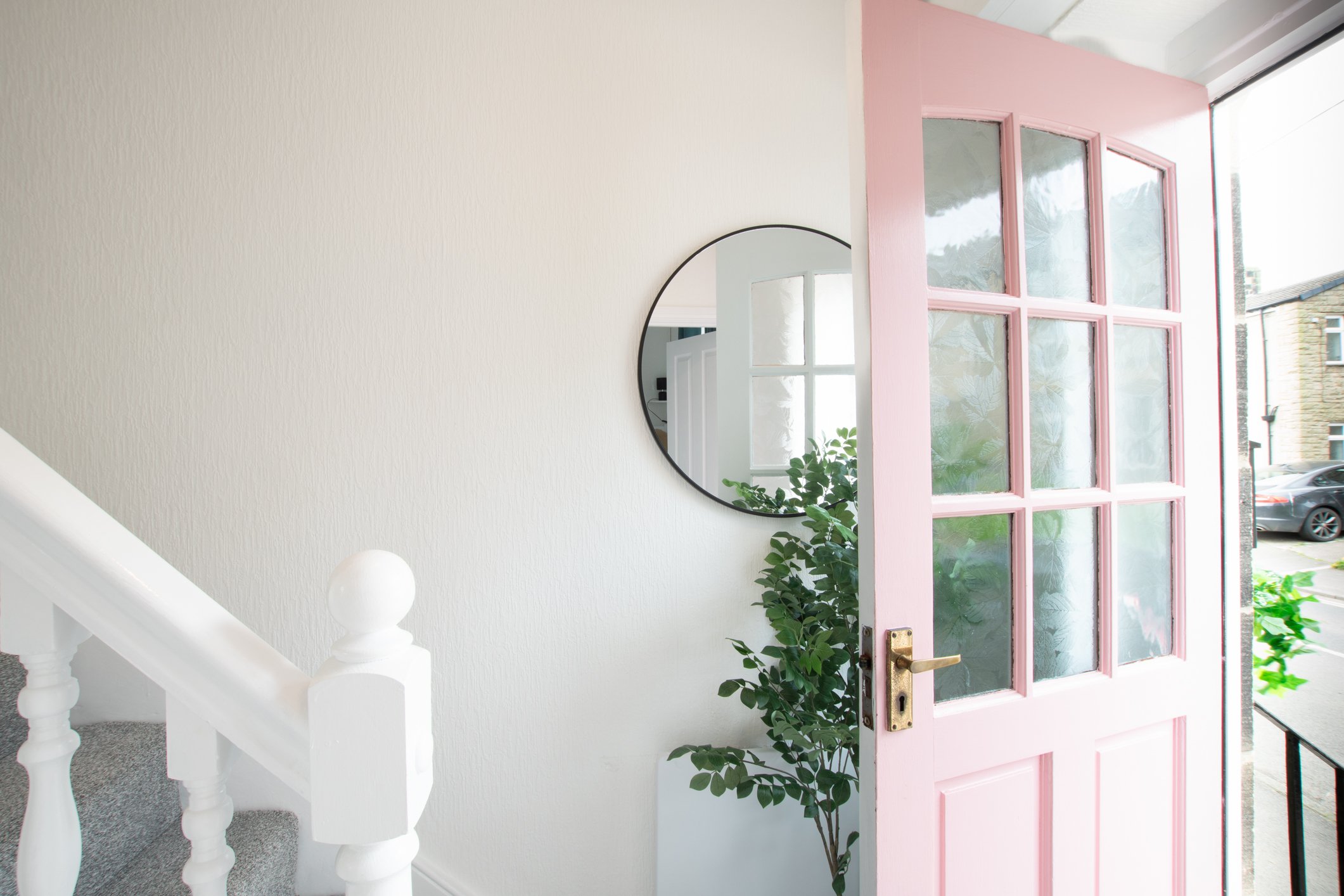Pink door with multiple glass panes, slightly open, showing a street view outside with a car parked nearby, green plants inside, white wall, mirror, and staircase inside the house.